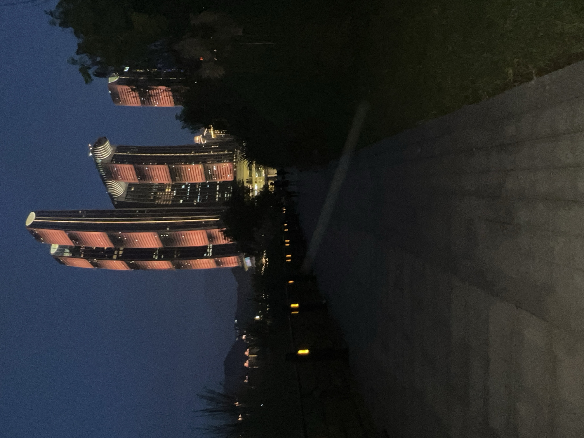 The image depicts a nighttime scene in Jiangbei Cheng Street, China. In the background, several modern skyscrapers are illuminated with red and white lights, creating a striking urban landscape against the dark blue sky. The buildings have unique architectural designs with segmented facades. In the foreground, a paved walkway runs through a park-like setting with trees and grass on the right side. Along the left edge of the walkway, there are decorative bollard lights with yellow illumination. A few figures are visible walking on the path in the middle ground, appearing as silhouettes against the lit buildings and walkway lights. The overall atmosphere is one of urban development and leisure at dusk.
