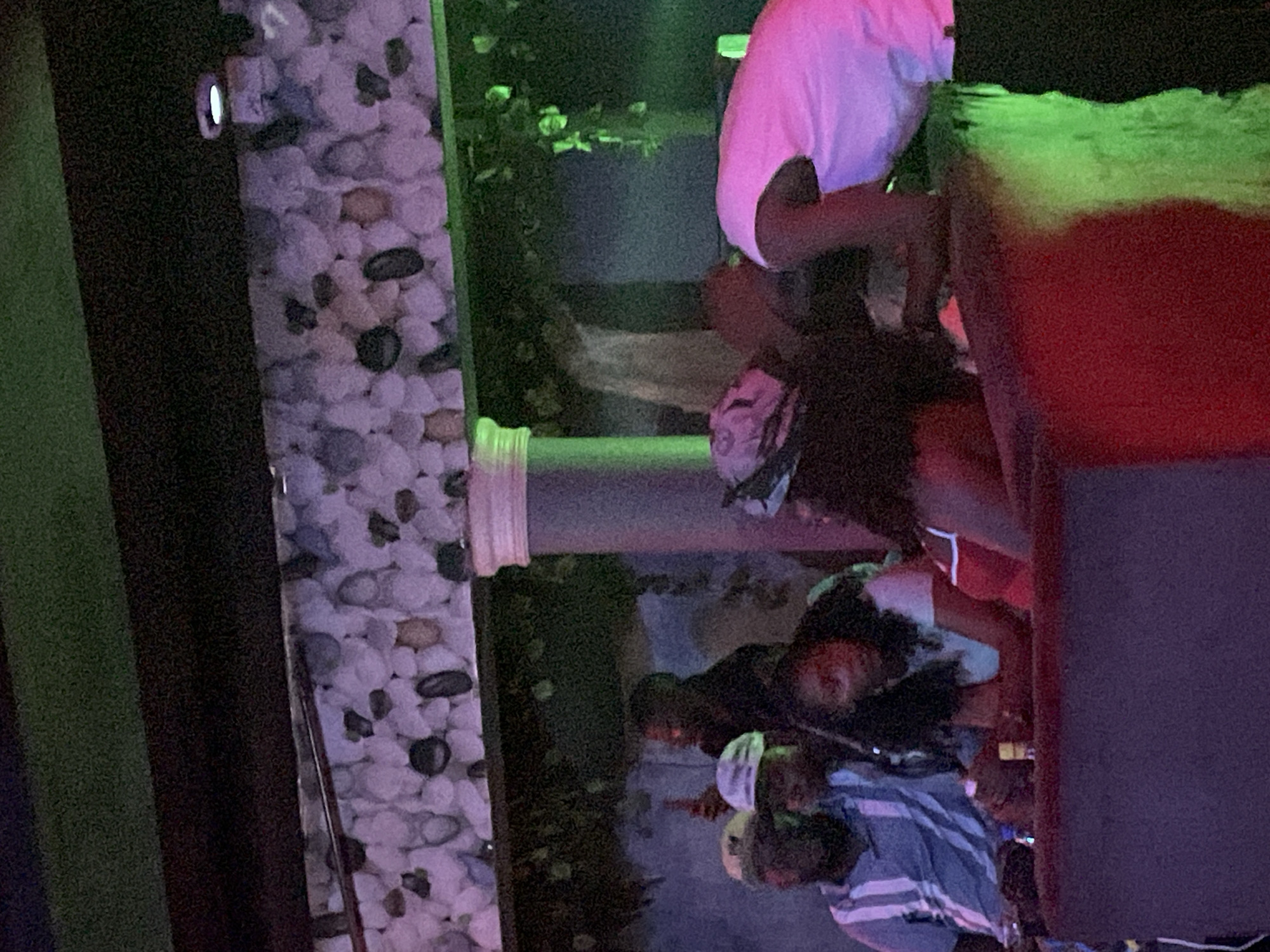 In the city of Durumi, Nigeria, a dimly lit establishment features patrons seated at a raised, red-upholstered counter. The background is adorned with a textured wall composed of light-colored, irregular stones, and overhead, dark beams are illuminated by green lighting. To the left, three men are visible; one wears a blue and white striped shirt and a light-colored cap. Adjacent to him, another man in a similar cap is partially obscured. Further back, a man in a dark shirt holds a bottle. In the center, a woman with dark, curly hair wears a light-colored shirt and looks down at her hands, which rest on the counter. Beside her, another woman, wearing a red top with white trim, is also seated. On the far right, a person in a pink top leans forward. Green lighting casts a glow on the right side of the scene.