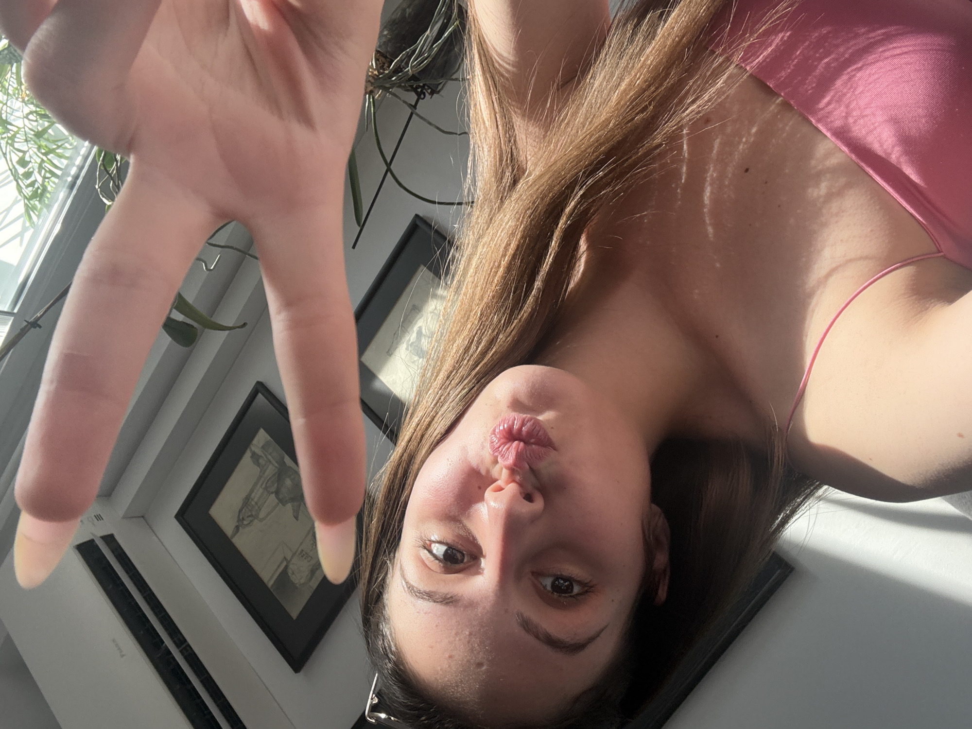 A young woman with long brown hair and a pink camisole is taking a selfie. She is making a duck face and holding up two fingers in a peace sign. The background includes a window with sunlight streaming through, a plant, and what appears to be an air conditioning unit. Two framed pictures are visible on the wall. The lighting suggests it is daytime. This scene is not identifiable as being in Burgas, Bulgaria, or any other specific city.