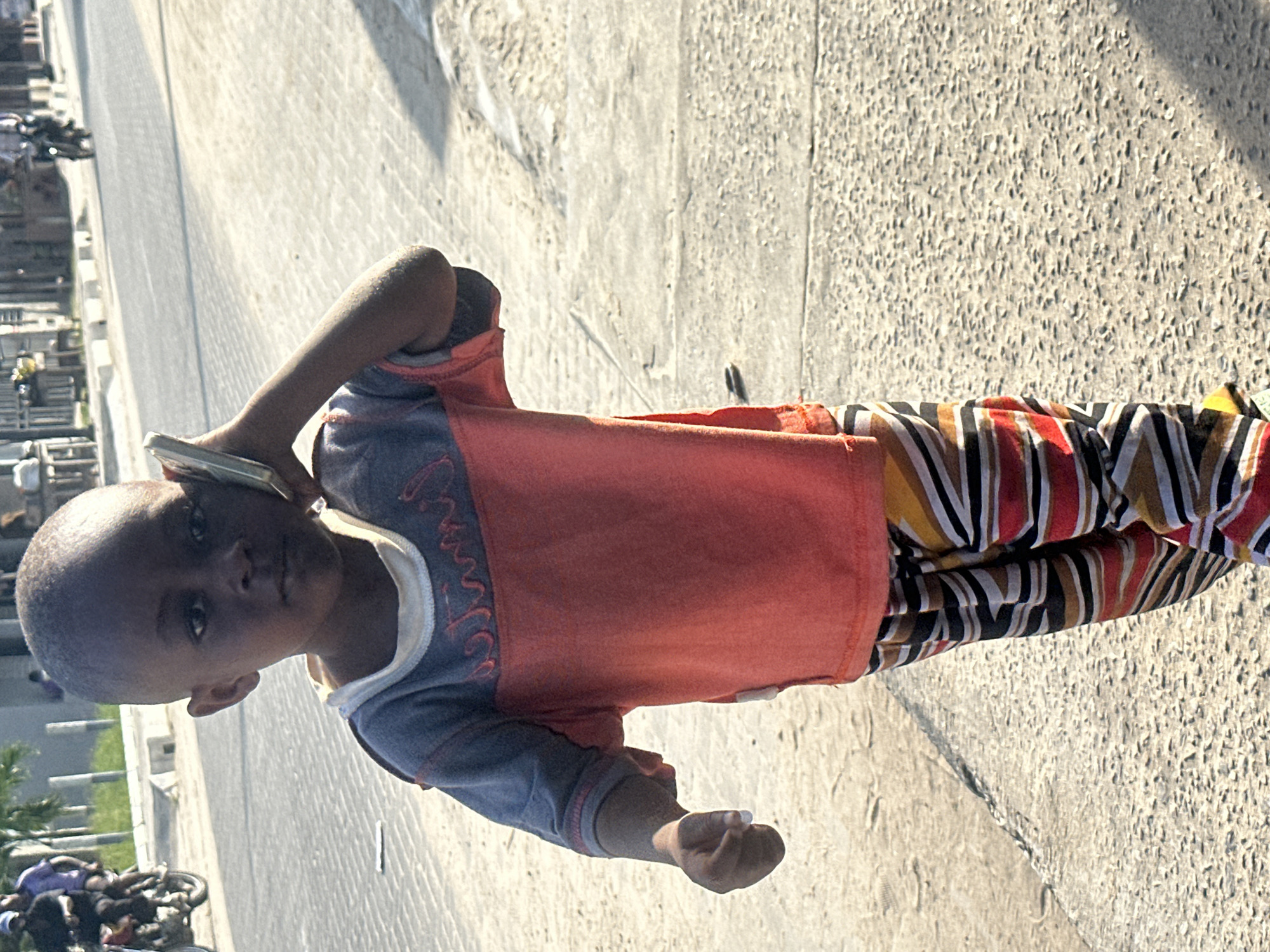 A young Black child, appearing to be between five and seven years old, stands on a paved road, holding a light-colored mobile phone to their right ear, as if in a conversation. The child is wearing a two-toned shirt, with the upper portion in a muted blue and the lower portion in a vibrant orange. Below the shirt, they are wearing striped pants featuring horizontal bands of red, yellow, white, and black. The child's left hand is slightly extended, with fingers partially open, holding a small white object, possibly a piece of paper or a small stone.
The child is positioned on the side of a road in what is identified as Badagry, Nigeria. The road surface is paved with interlocking blocks. In the background, to the left, a person on a motorcycle is visible, wearing a purple shirt and a helmet. Further in the background, more structures and people are discernible, with trees and possibly some buildings on the left side of the frame. The lighting suggests it is daytime, with shadows cast on the ground. The overall environment appears to be an urban or semi-urban street scene.