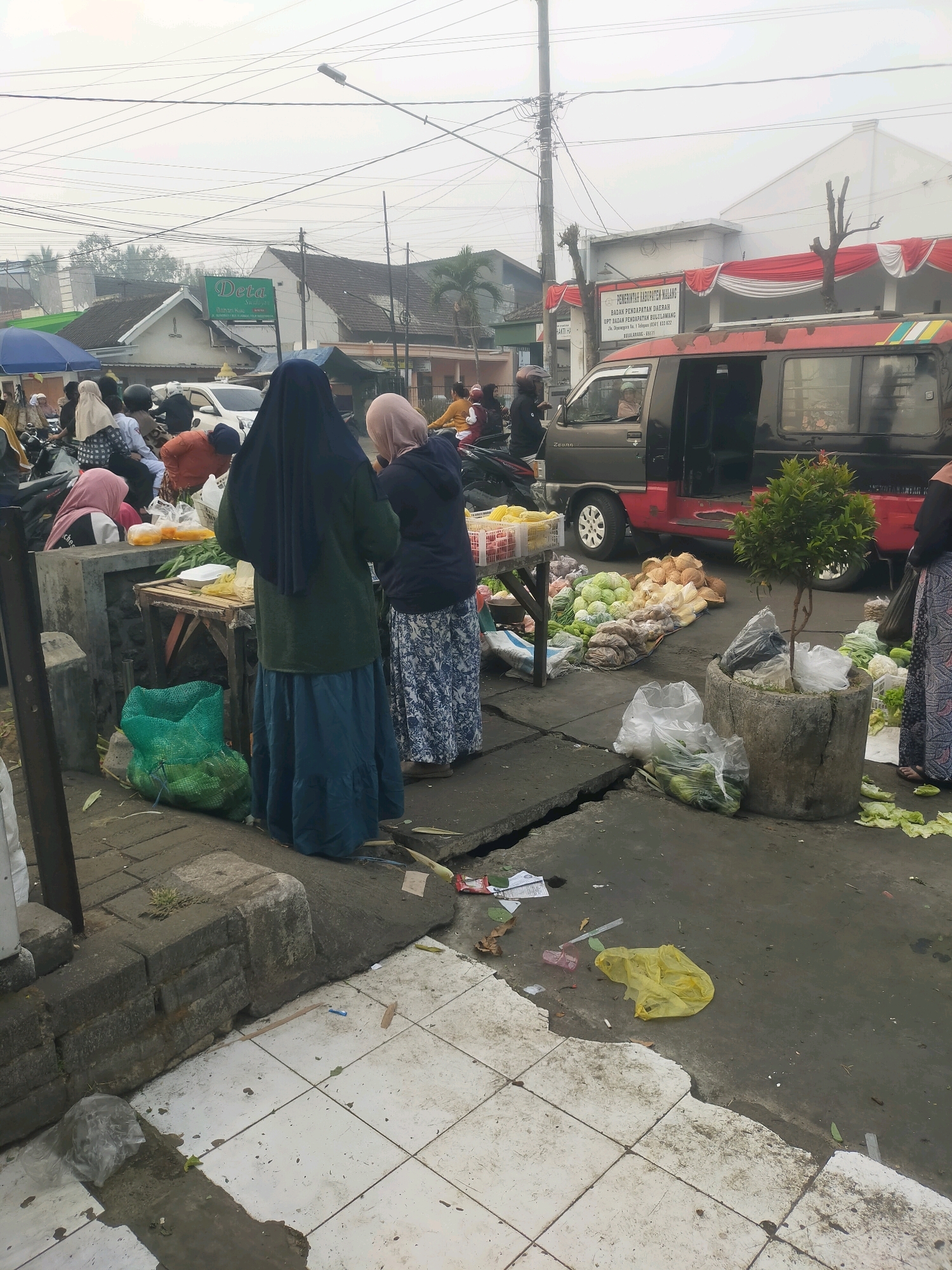 The image depicts a street scene in Bululawang, Indonesia, likely a market area given the presence of vendors and produce. In the foreground, a woman wearing a dark blue hijab and a green jacket stands next to a table laden with food items. Behind her, another woman in a light pink hijab is visible. To the right, a concrete planter contains a small tree, and various vegetables like cabbage and coconuts are displayed on the ground. A red and black van is parked nearby, and several motorcycles are scattered throughout the scene, indicating a busy urban environment. The overall atmosphere suggests a typical day at a local market, with people engaged in commerce and daily activities.