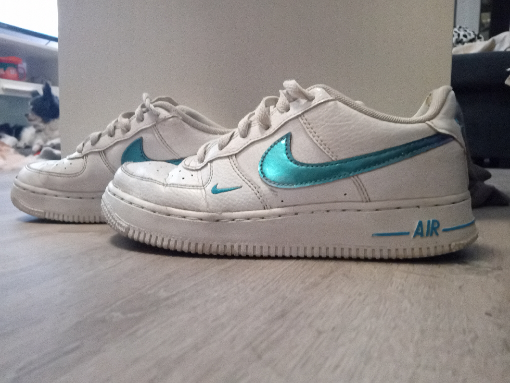 Two white Nike Air Force 1 shoes with teal accents rest on a light wood-colored floor. The laces are tied and the shoes appear slightly worn. In the background, a blurred area suggests a domestic interior with a shelf, possibly a television, and a dog visible. The room is indoors, and the lighting is soft. The phrase "AIR" is written on the side of the closest shoe in teal.