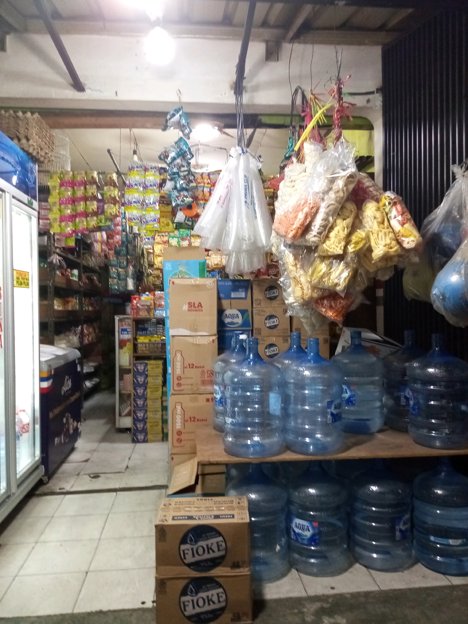 The image depicts the interior of a small convenience store, likely a warung, located in Bekasi, Indonesia. The store appears well-stocked with a variety of goods. In the foreground, a wooden shelf holds several large, clear plastic water jugs, some bearing the "AQUA" logo. Below this shelf, more water jugs are arranged. To the left, a glass-doored refrigerator or cooler is visible, containing bottled drinks.
Behind the water jugs and refrigerator, the store's shelves are packed with numerous packaged goods. These include snacks in colorful wrappers hanging from the ceiling, as well as boxes and bags of food items stacked on shelves. Prominently displayed are boxes of "FIOKE" branded products. The store is lit by overhead fluorescent lights, creating a bright and somewhat uneven illumination. The floor is tiled. The overall impression is of a busy, albeit cluttered, retail space offering everyday necessities.