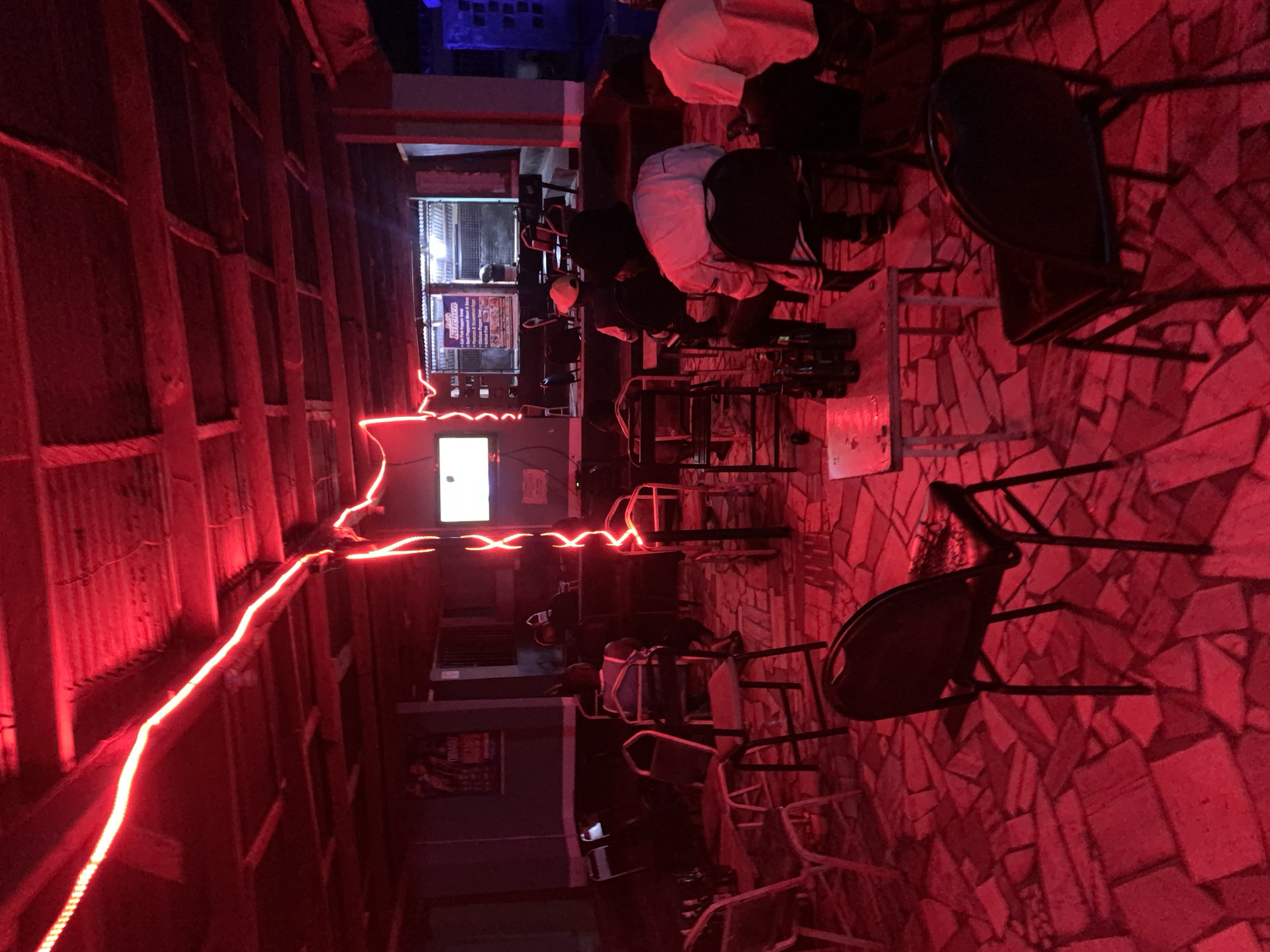 The scene is a dimly lit bar interior, possibly in the vicinity of New Stadium, Nigeria, judging by the building materials and interior design. Red string lights illuminate a corrugated metal and wooden structure forming a partial ceiling or wall. Several patrons are seated at bar stools, their backs to the camera. A bar counter is visible with equipment and signage. The floor appears to be constructed of irregular stone tiles, also heavily illuminated in red. The focus is on the interplay of light and shadow, and the social environment within the bar.