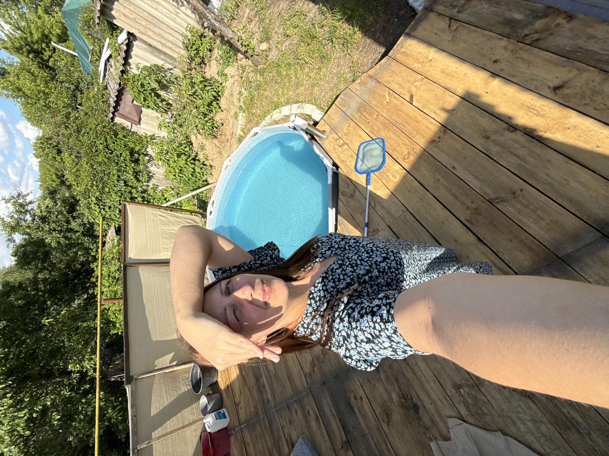A woman is lying on a wooden deck next to a small, blue inflatable pool. She is wearing a dark blue floral print dress and has her hand on her forehead, as if shielding her eyes from the sun. The sun is casting long shadows across the deck, indicating it is likely late afternoon or early evening. The scene is outdoors, with lush green trees and a cloudy sky visible in the background. A blue pool skimmer rests on the deck near the pool. To the left of the woman are several objects, including buckets and a white plastic container. The overall mood appears relaxed and summery.