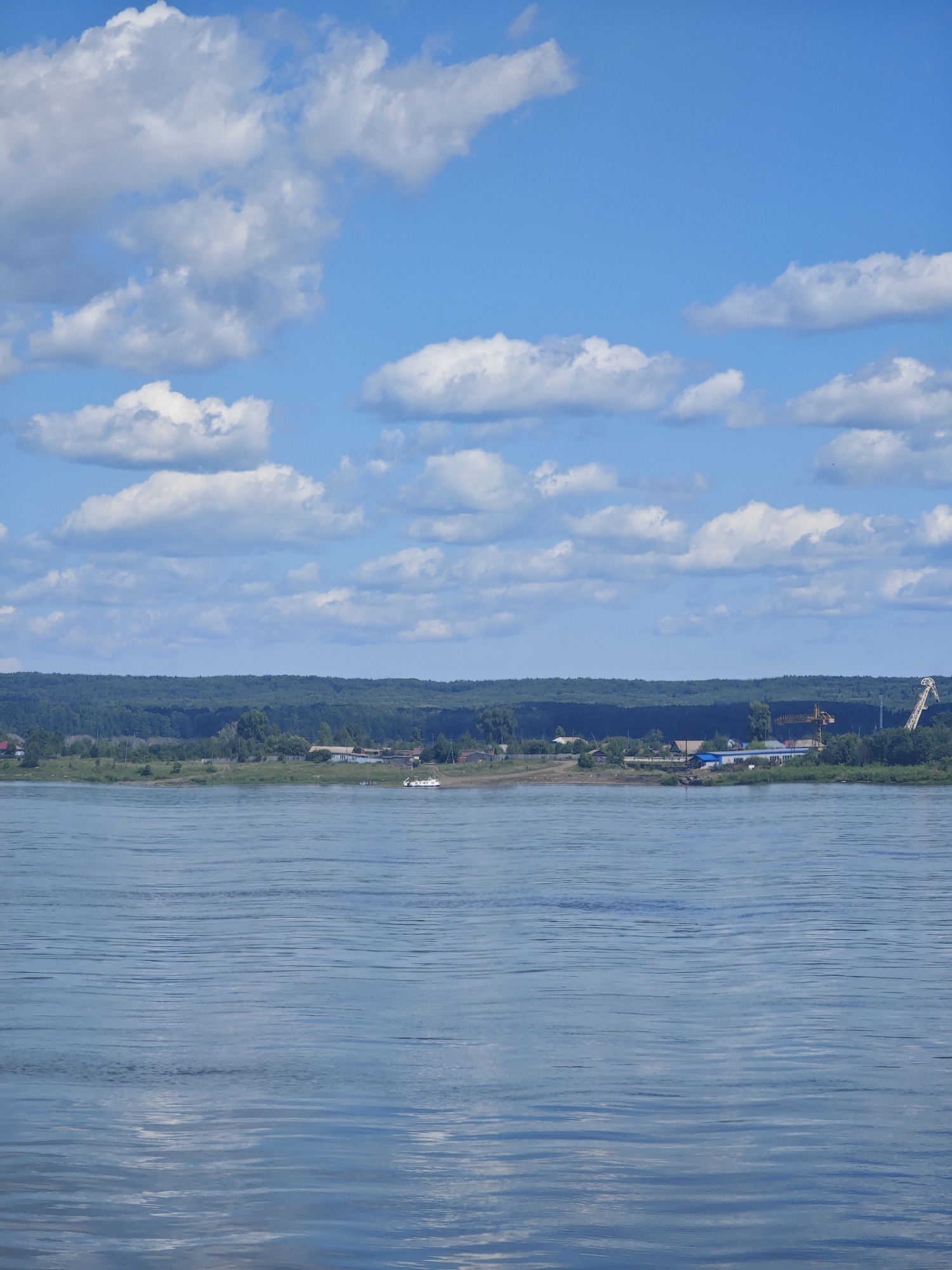 An aerial view shows a wide river under a bright blue sky with scattered white clouds. On the far bank, a dense green forest stretches across the horizon. In front of the forest, a strip of land hosts a small settlement with various buildings and structures, including a yellow construction crane on the right side of the frame and a white boat docked near the shore. The river's surface is rippled, reflecting the sky and clouds. The location is the city of Strelka, Russia.