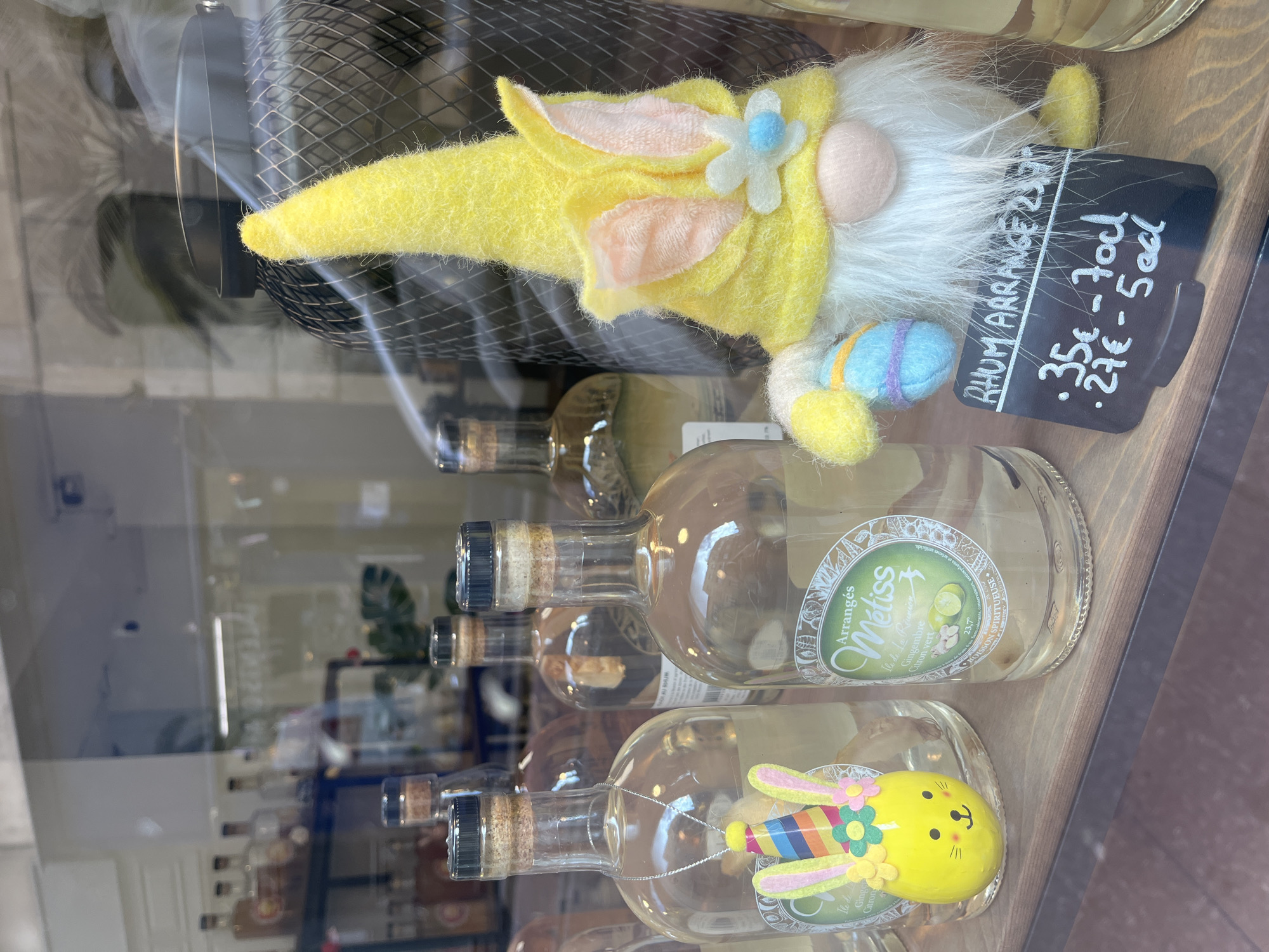 The image captures a display in the city of Metz, France, featuring bottles of "Arrangés Métiss" rum, each adorned with different Easter-themed decorations. Some bottles are empty, and some have Easter decorations inside. The rum bottles are labeled with a sticker describing their flavor profile as “Ile de Gingembre Citron Vert” and include a small white label advertising a small bottle as “27€ -50cl” or the large as "35€ -70cl". At the top right is a yellow gnome figure holding a miniature Easter egg. The gnome has a white beard and a pink nose. The display is situated behind a glass window, reflecting the street scene outside.