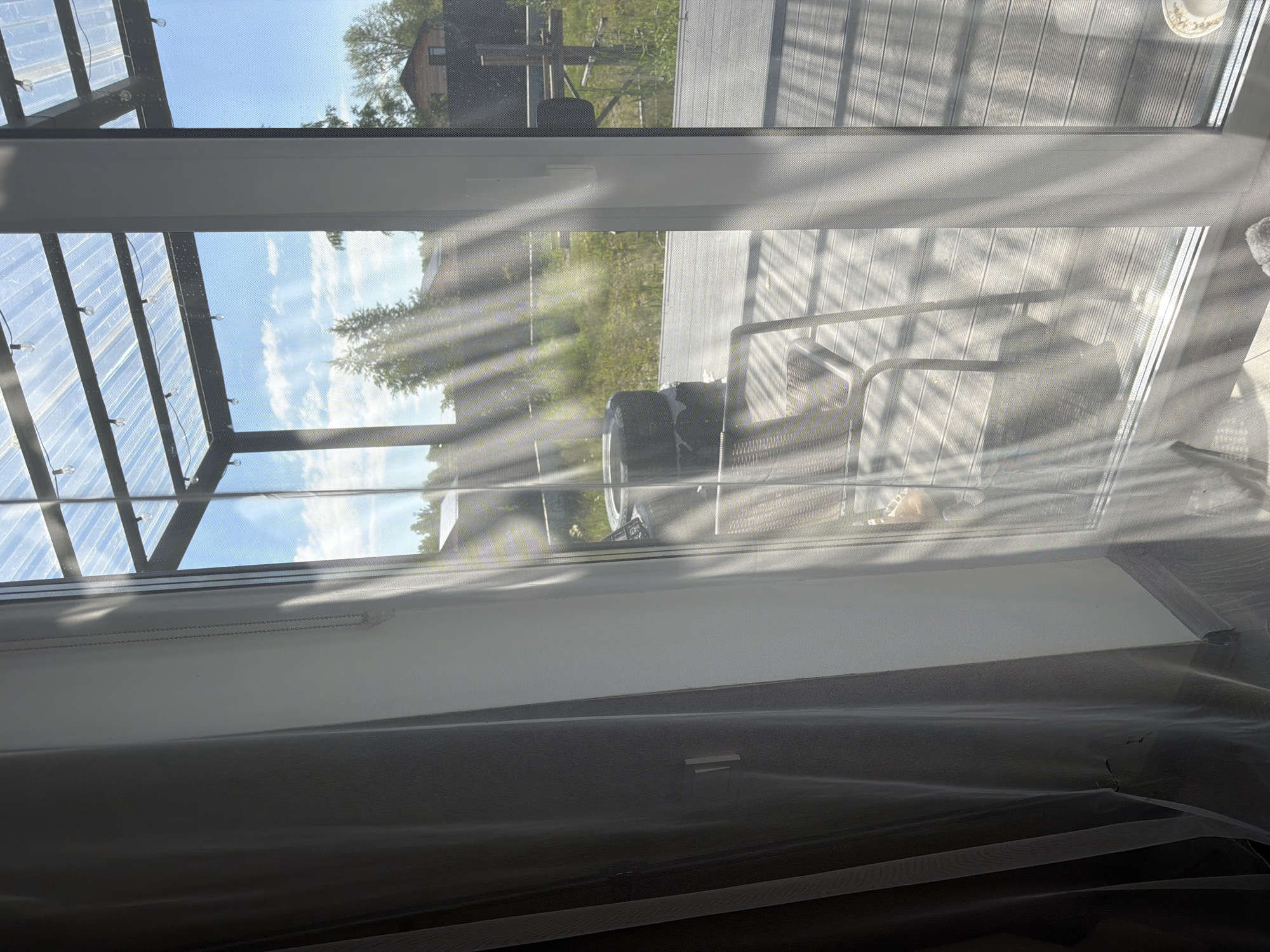 The image depicts an outdoor patio area with a wooden deck, viewed through a glass door and sheer curtains. Sunlight streams through a translucent roof structure, casting diagonal shadows across the scene. On the deck, a metal chair and a stack of tires are visible. In the background, there is a grassy area with some trees and a building. The overall impression is one of a residential backyard on a sunny day. The location is identified as Malinovka, Russia.