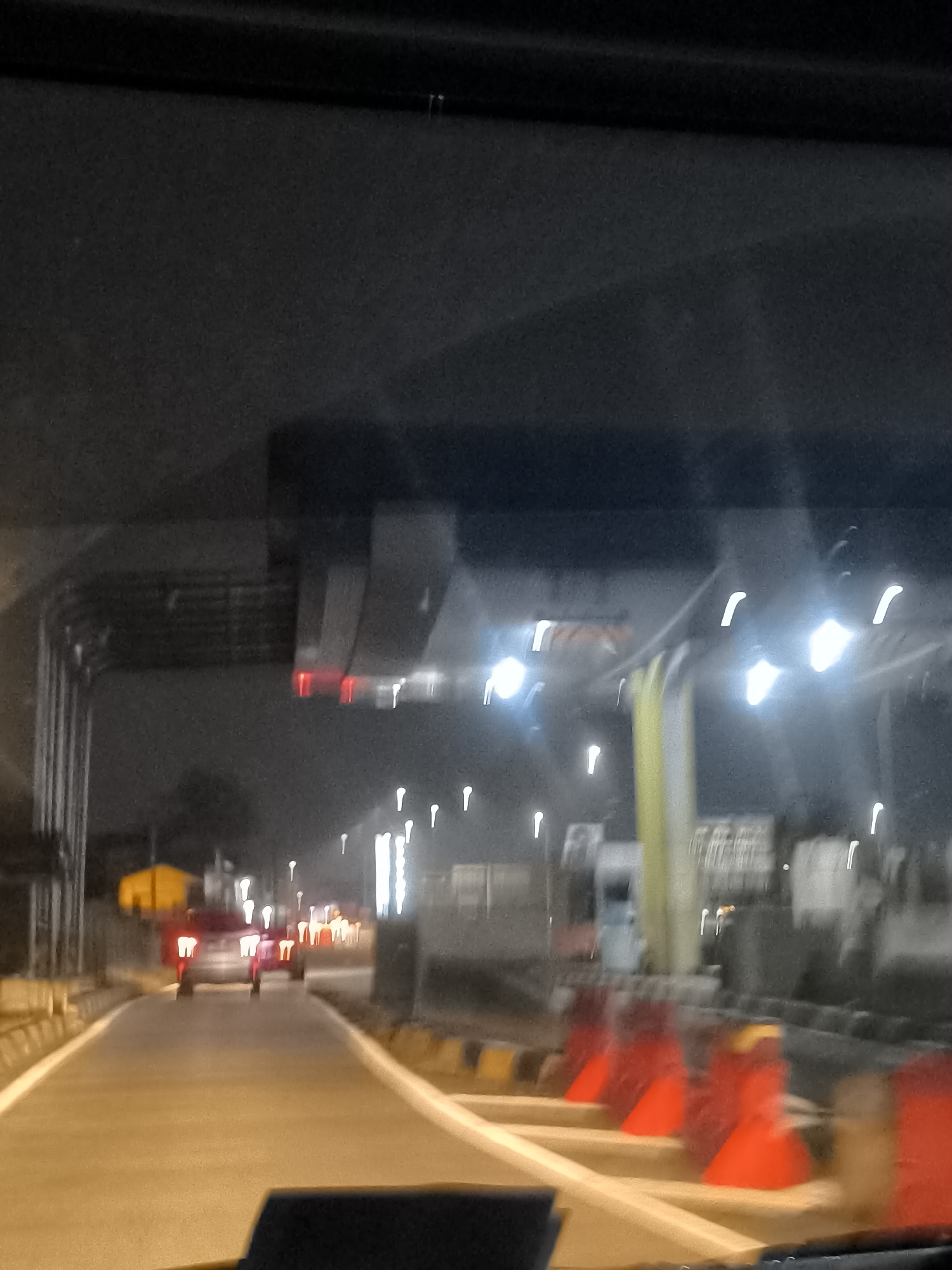 The image depicts a nighttime view of a toll gate in Depok, Indonesia, likely on the Jagorawi Toll Road or a similar arterial route. The perspective is from inside a vehicle, suggesting the photographer is either driving or a passenger. The toll gate structure is visible in the background, with illuminated signs and barriers. Several vehicles, including cars and possibly a truck or bus, are present at the toll booths, their headlights and taillights creating streaks of light due to the motion of the camera. The road surface is asphalt, and there are traffic cones and safety barriers lining the lanes. The overall atmosphere is dimly lit, characteristic of a highway at night, with artificial lighting from the toll gate and vehicle headlights dominating the scene. The blurriness of the image indicates movement, likely from the vehicle from which the photo was taken.
