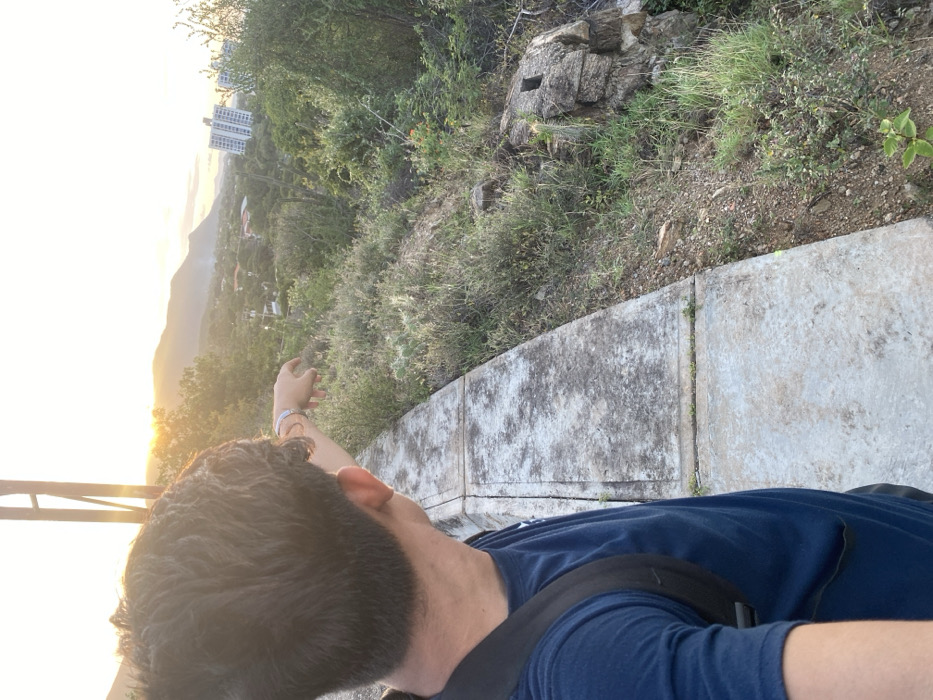 A person with dark hair and a blue shirt stands on a concrete ledge, facing a scenic view. In the background, hills stretch towards the horizon, bathed in the warm glow of sunlight. Trees and bushes line the edge, transitioning into a rocky area further back. The picture appears to be taken from a high vantage point, possibly a hike.
