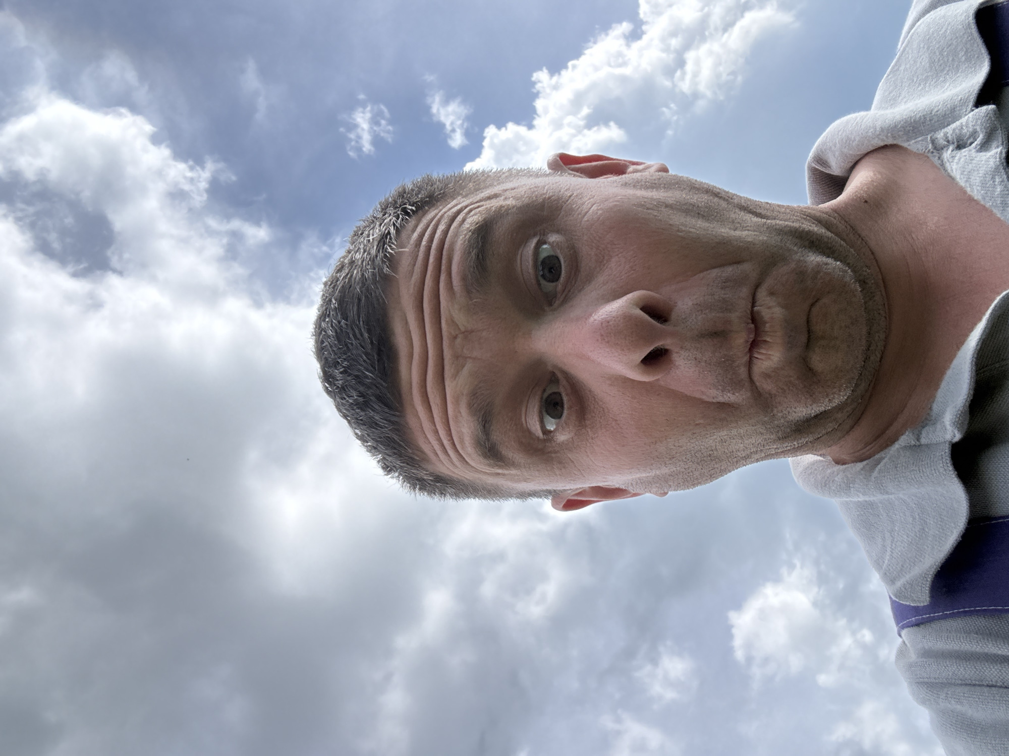 The media file is a low-angle selfie of a person against a cloudy sky. The person is leaning in towards the camera, with their face filling the majority of the frame. They have short, graying hair and visible wrinkles on their forehead. Their eyes are open and looking directly at the camera with a surprised or startled expression. Their mouth is slightly open. They are wearing a light gray hooded garment with purple accents. The background is a sky filled with dramatic, gray and white clouds, suggesting overcast weather. The time of day is not explicitly indicated, but the lighting is diffused, consistent with daytime under cloud cover. There is no visible text in the image. The perspective is from below, looking up at the person, and the image is oriented with the person's head at the bottom of the frame. The setting appears to be outdoors.