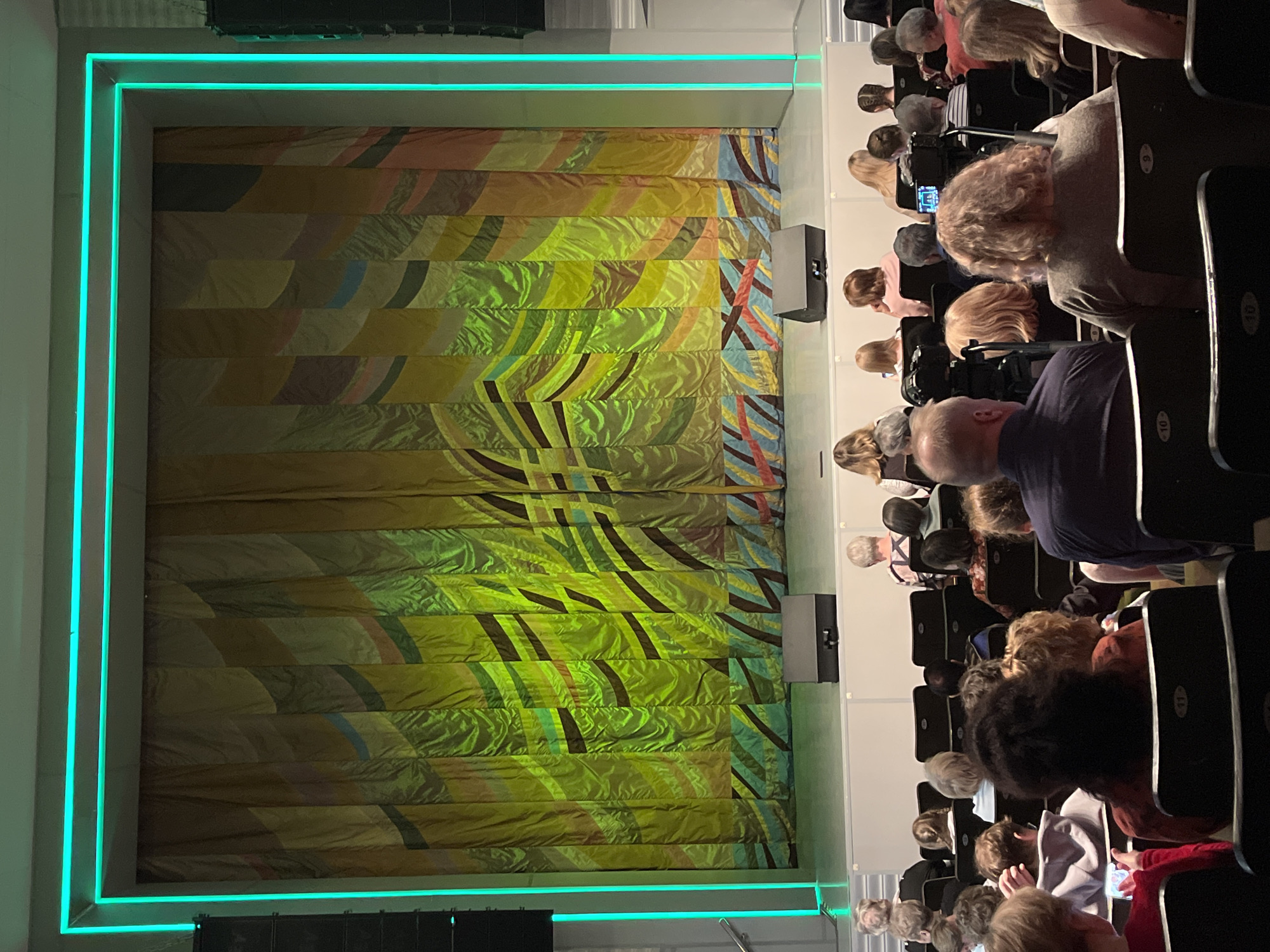 An image depicting an audience seated in a theater. The audience is facing a stage adorned with a vibrant, patterned curtain illuminated by neon lights. The location is presumed to be in Chaikovsky, Russia, based on the context of the prompt. The audience consists of a diverse group of individuals, primarily adults, seated in rows of black chairs, each numbered. The focus is on the backs of their heads and shoulders. The stage curtain features a design of horizontal stripes, alternating between shades of yellow, green, brown, and other colors, with abstract patterns. A neon light frame surrounds the curtain, casting a blue-green glow. Black speakers are visible at the top and bottom.