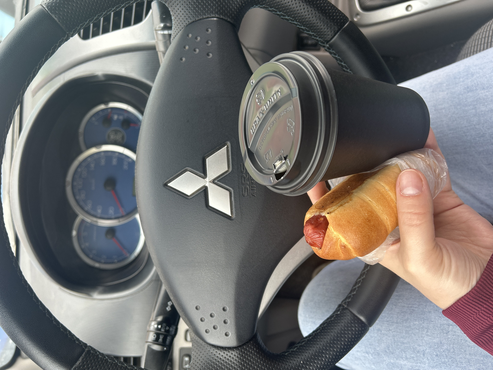 The image depicts the interior of a Mitsubishi vehicle, specifically in the city of Chaikovsky, Russia. A person, whose lower torso and left hand are visible, is seated in what appears to be the driver's seat. The person is wearing light blue jeans and a burgundy long-sleeved top. The left hand holds a hot dog encased in a clear plastic bag and a black disposable coffee cup.
The hot dog consists of a reddish sausage in a light-brown bun, with visible ketchup on top of the sausage. The black coffee cup has a dark, possibly camouflage-patterned lid. Visible text on the lid includes Cyrillic script reading "УРА! ХОРОШИЙ!" (Ura! Khoroshyi! - Hooray! Good!) and "ОСТОРОЖНО ГОРЯЧО!" (Ostorozhno Goryacho! - Caution Hot!), along with partially obscured English text "UP2U QUALITY". A small opening for sipping is present on the lid.
The steering wheel, centered in the frame, is black, leather-wrapped, and features a silver Mitsubishi logo. Below the logo, "SRS AIRBAG" is inscribed. The steering wheel has visible blue-green stitching on the right side and a perforated grip section. Controls are partially visible on the left spoke of the steering wheel. Behind the steering wheel, the instrument cluster with two prominent blue-faced gauges and red needles is visible. To the left, a multi-function stalk for turn signals and lights is visible. The dashboard is predominantly dark grey/black.
