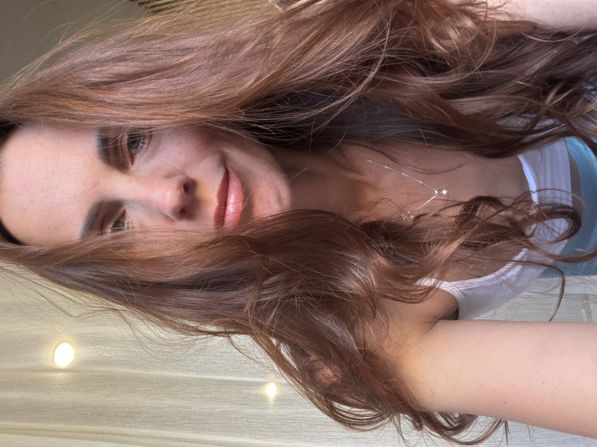 This close-up portrait features a person, likely female, with long, wavy reddish-brown hair. The image is taken from an overhead perspective, with the subject looking up towards the camera while seemingly lying down or leaning back. The photo itself is rotated 90 degrees clockwise.
The subject has light skin, light-colored eyes (appearing grey-blue or green), and a subtle, gentle smile on their full, pink-tinted lips. Their voluminous hair cascades around their head and shoulders, framing the face and spreading across the background surface. They are wearing a white tank top or camisole, with a glimpse of light blue fabric (possibly a bra strap or inner layer) visible on the right side. A delicate silver chain necklace with two small, sparkling pendants adorns their neck.
The background shows an indoor setting, characterized by a light-colored, possibly wooden or textured wall/floor panel. Two distinct circular light sources, resembling recessed spotlights, emit a warm glow in the background. The lighting is soft and ambient, creating a relaxed mood. The subject appears calm and content. No visible text is present in the image. The location context for this media file is Birobidzhan, Russia.