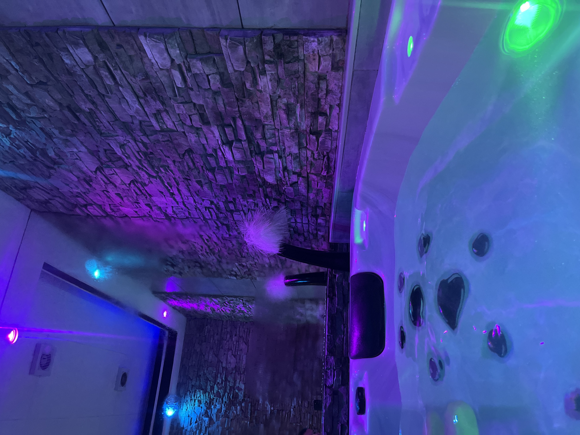 The image shows a detail of a spa or hot tub area. The walls are covered with a rough stone-like material illuminated with purple light. A white, fluffy object, possibly a decorative element, is attached to a dark-colored fixture near the wall. Part of the hot tub is visible on the right, lit with blue and green lights, showing the water's surface and several jet openings. A steam effect is present, contributing to the spa atmosphere. The image does not contain enough information to confidently pinpoint the location as Zagreb, Croatia.