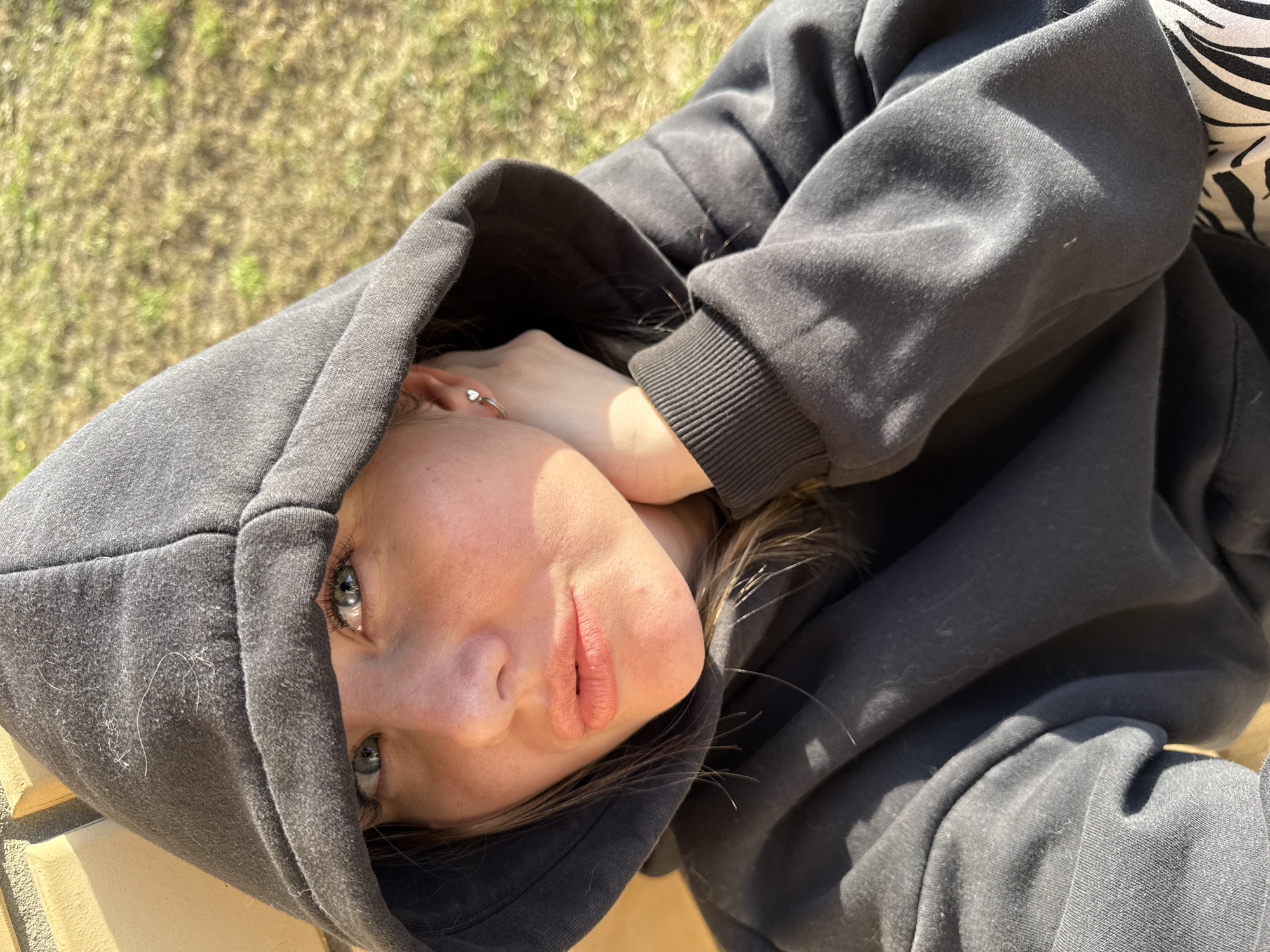 An overhead shot captures a young person, likely female, lying down and looking directly at the camera. Their head is tilted, and they are wearing a dark charcoal grey hooded sweatshirt (hoodie) with ribbed cuffs. A small, heart-shaped ring is visible on a finger of one hand, which is held near their ear.
The person has light skin, blue-grey eyes, and light brown hair partially visible around their face and beneath the hood. Their expression is neutral to slightly contemplative, with lips slightly parted. The fabric of the hoodie shows some lint or fibers, particularly on the hood.
In the upper right corner, a glimpse of a black and white zebra print pattern is visible, possibly from another piece of clothing or a blanket. The background is softly blurred, revealing dry, yellowish-green grass or foliage, suggesting an outdoor setting. Bright sunlight illuminates the scene, casting soft shadows, indicating it is daytime, possibly morning or late afternoon, under clear skies in Gornyi, Russia. There is no visible text in the image.