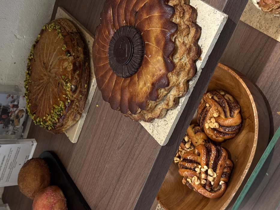 The image depicts a multi-tiered display counter inside a French bakery or pâtisserie in Paris, France. The shelves are made of a dark brown wood-grain material.
On the top shelf, to the left, two round, golden-brown pastries resembling brioche or large doughnuts, one with a reddish sugar coating, rest on a matte black rectangular tray. To their right, a large, round Galette des Rois with a golden, patterned crust is presented on a light beige, square stone tile, its surface dotted with chopped green pistachios. Behind these items, a clear acrylic sign displays French text, including the prominent phrase "GALETTE DES REINES."
The middle shelf features another large, round Galette des Rois, also situated on a light beige stone tile. This pastry has a deeply browned, fluted edge and is adorned with a central dark chocolate medallion bearing an embossed design.
Partially visible on the bottom shelf, within a natural wood bowl, are three swirled, golden-brown pastries infused with a dark filling and topped with chopped hazelnuts. A faint green reflection, likely from a display case glass, is present at the bottom edge of the frame. No individuals are present in the scene.