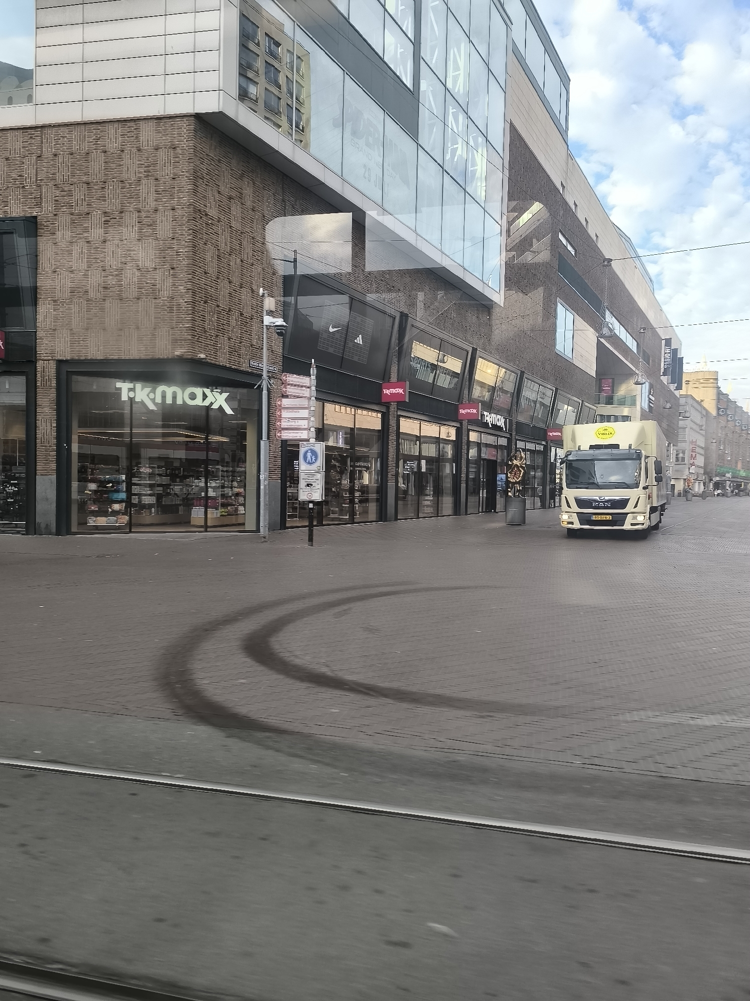 The image depicts a street scene in The Hague, Netherlands, likely a commercial area. In the foreground, tram tracks run across the paved street, with tire marks from a vehicle visible on the cobblestones. A modern building with a brick facade and large glass windows dominates the left side of the frame. The TK Maxx logo is prominently displayed on the storefront. Further down the street, a beige delivery truck with the logo "VELTA" on its roof is stationary. Several other storefronts, some with "TK Maxx" signage, are visible along the building's facade. The sky is cloudy. The overall scene suggests a typical urban environment with retail establishments and street infrastructure.