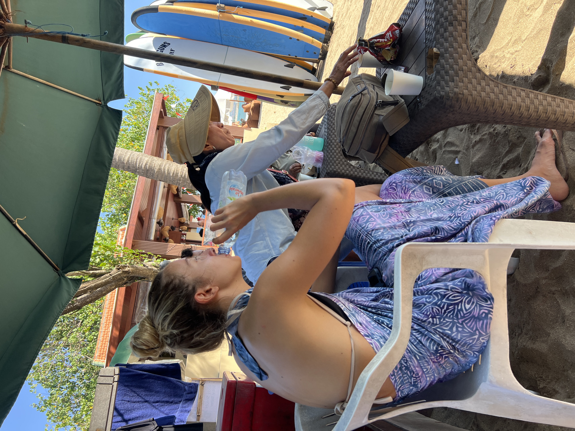 Two women are sitting at a table on a beach in Kuta, Indonesia. The woman on the left, with blonde hair in a bun, is wearing a purple and white patterned sarong and drinking from a plastic water bottle. The woman on the right, wearing a tan sun hat and a light-colored shirt, is reaching for something on the table. The table is made of wicker and has two white cups, a bag, and a packet of chips on it. Behind the women, several surfboards are standing upright. The scene is shaded by a large green umbrella, and in the background, there are trees, buildings, and the ocean. The sand is visible on the ground.