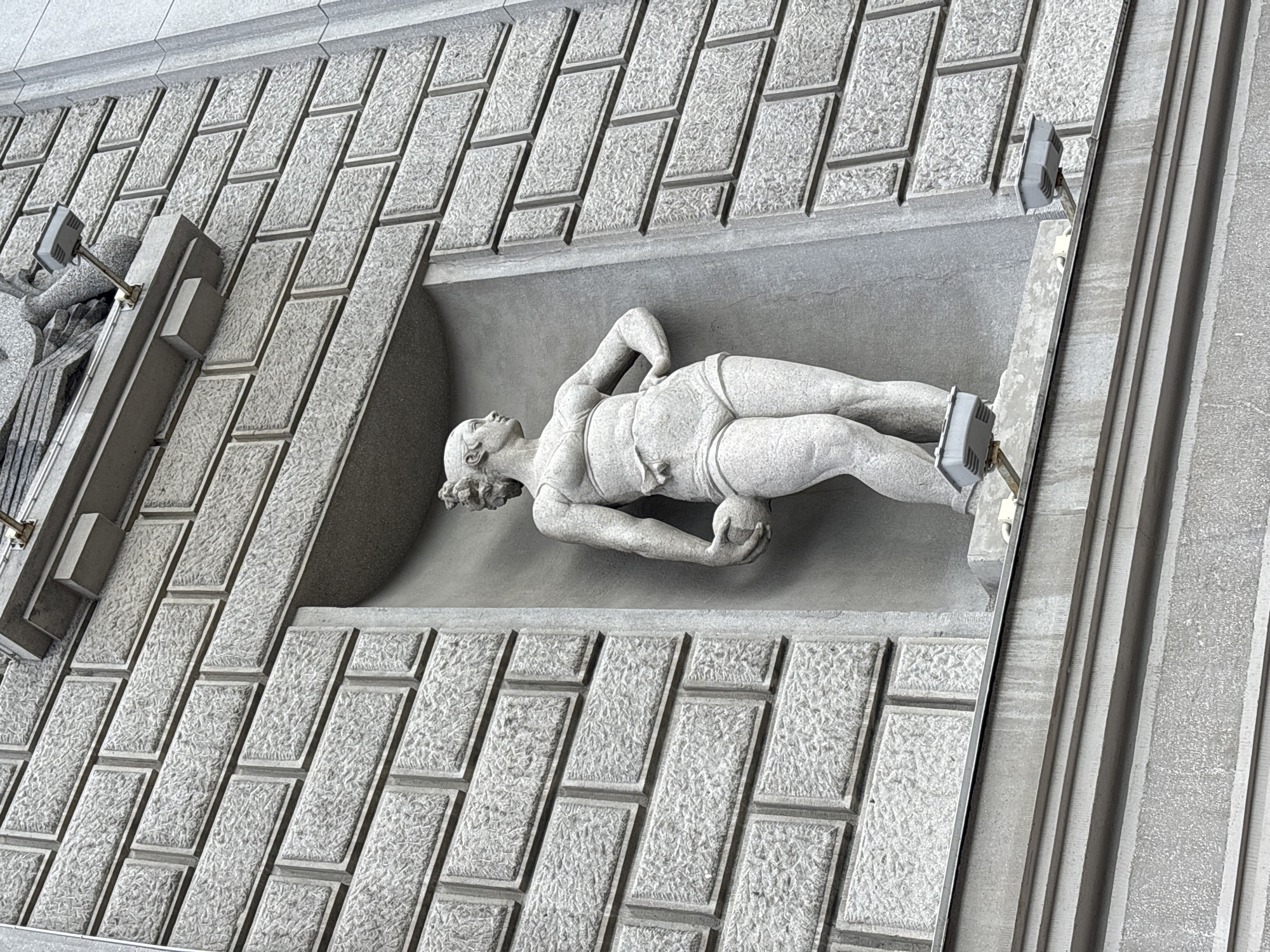 A low-angle, full-shot photograph captures a gray stone sculpture of a female figure standing in a recessed niche on a stone-clad building in Novokuybyshevsk, Russia. The sculpture depicts a muscular woman with her hair pulled back in a bun, wearing a brief athletic outfit. She stands with her left hand on her hip and her right arm bent, holding a spherical object against her side. The sculpture is illuminated by two gray, box-shaped lights mounted on brackets on the building's facade below the niche. The building's exterior is constructed of large, gray stone blocks arranged in a brick pattern. A portion of a carved, winged figure is visible at the top of the frame, suggesting it is part of a larger architectural embellishment. The overall tone of the image is muted and monochromatic, emphasizing the textures of the stone.