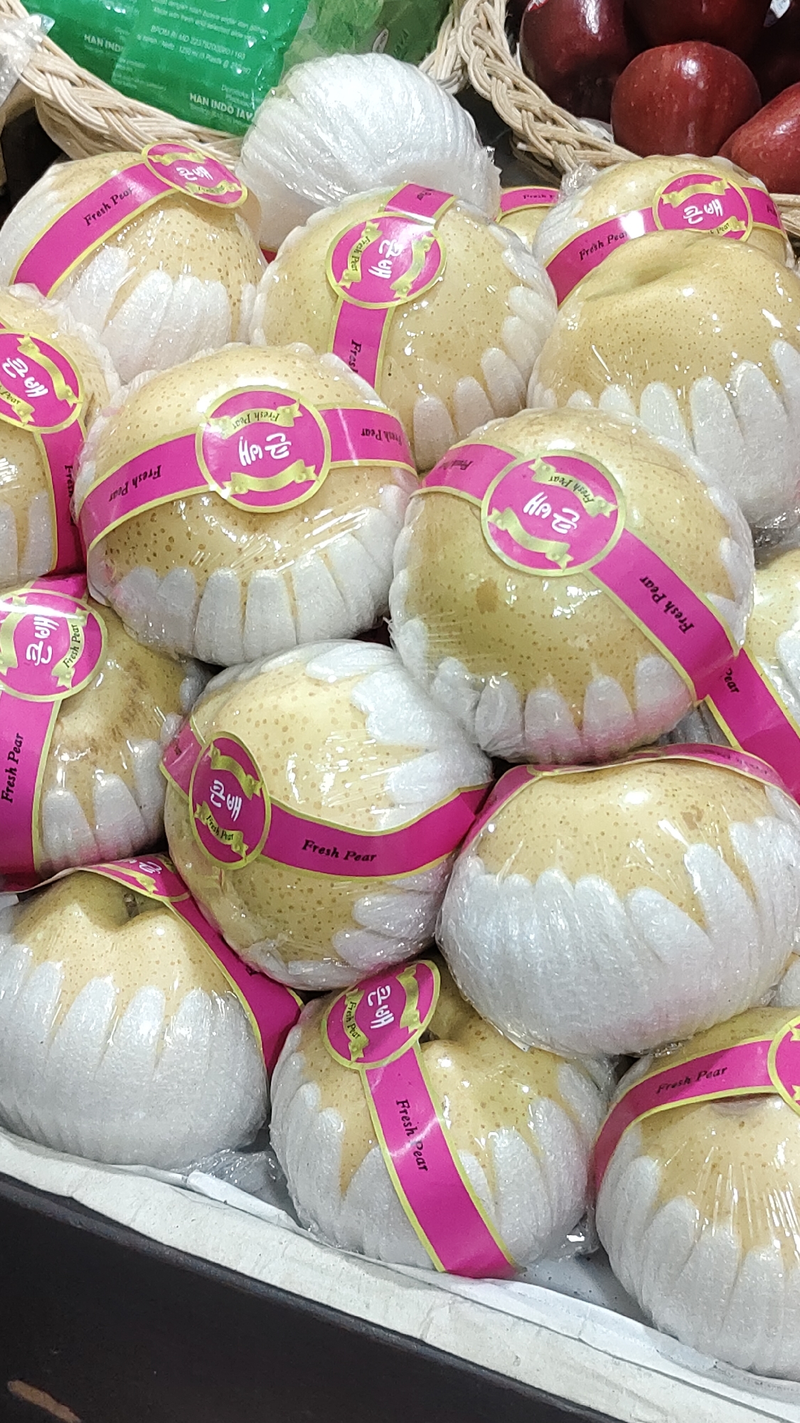 The image shows a close-up, eye-level view of a stack of individually wrapped fresh pears. The pears are light yellow and are encased in a clear plastic wrap, with a white foamy material at the bottom of each pear. A bright pink band with yellow text wraps around the middle of each pear. The text on the pink bands appears to be in Korean and English, with the English text clearly reading "Fresh Pear". Some of the Korean characters are visible. The pears are arranged in a basket-like container, and in the background, a green plastic bag with some text and red apples are partially visible. The lighting is bright, suggesting an indoor setting, likely a market or grocery store. There are no people or animals visible in the image. The location is identified as a market in Malang, Indonesia.
