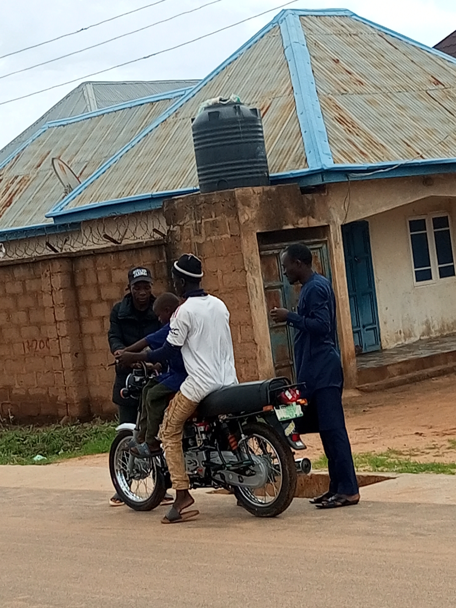 In Tempure, Nigeria, three individuals are gathered around a motorcycle parked on a dirt road. The first person, wearing a black jacket and a black baseball cap, is standing to the left of the motorcycle. The second person, a child wearing a blue shirt and green pants, is seated on the motorcycle. The third person, wearing a white t-shirt and tan pants, is also seated on the motorcycle behind the child. A fourth individual, dressed in a dark blue traditional outfit, stands to the right of the motorcycle, appearing to interact with the people on it. In the background, a building with a corrugated metal roof and blue trim is visible, along with a water tank and a brick wall. The sky is overcast.
