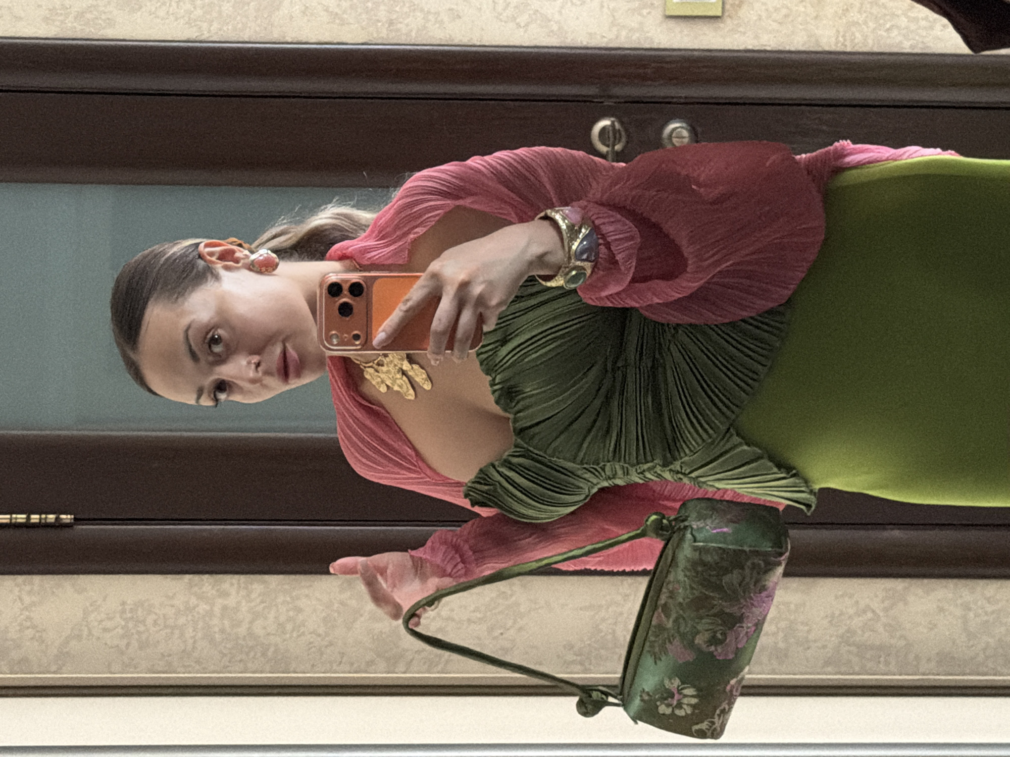 A woman stands in front of a mirror, taking a selfie. She is wearing a green, ruched dress and a pink shawl. She is also carrying a green floral-patterned handbag and wearing statement jewelry, including a gold necklace, large earrings, and a bracelet. The background appears to be an indoor setting, possibly a hallway or entranceway, with wooden doors and textured walls. There are no overt indicators of the location being specifically Krasnoyarsk, Russia, beyond the prompt's assertion.