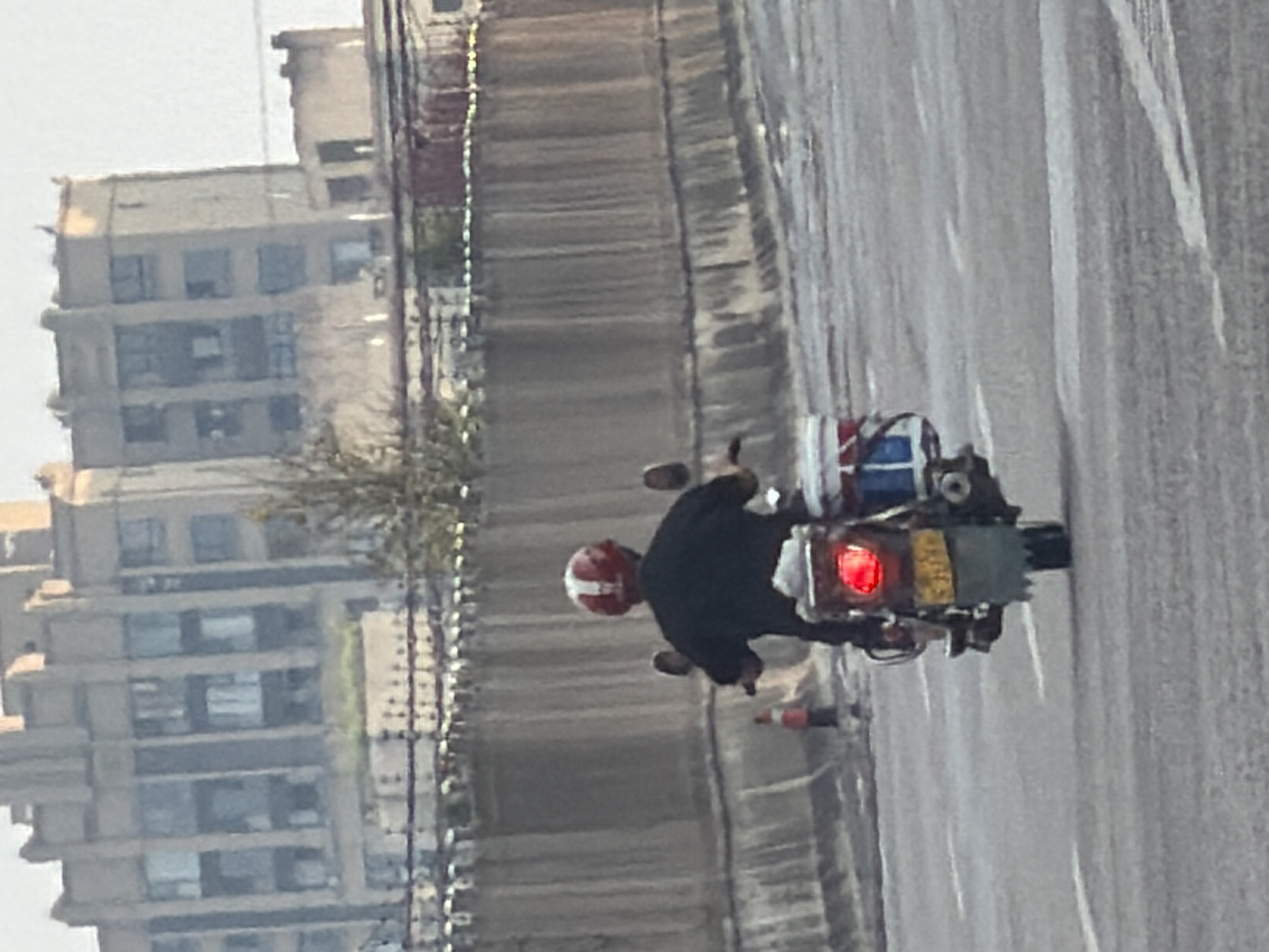 A person wearing a black shirt and a red and white helmet is riding a motorcycle on a paved road. The motorcycle has a red taillight and a yellow license plate. There is a tall building in the background and a wall with a decorative fence. The scene is likely in the city of Jiangjin, China, as indicated by the license plate style.