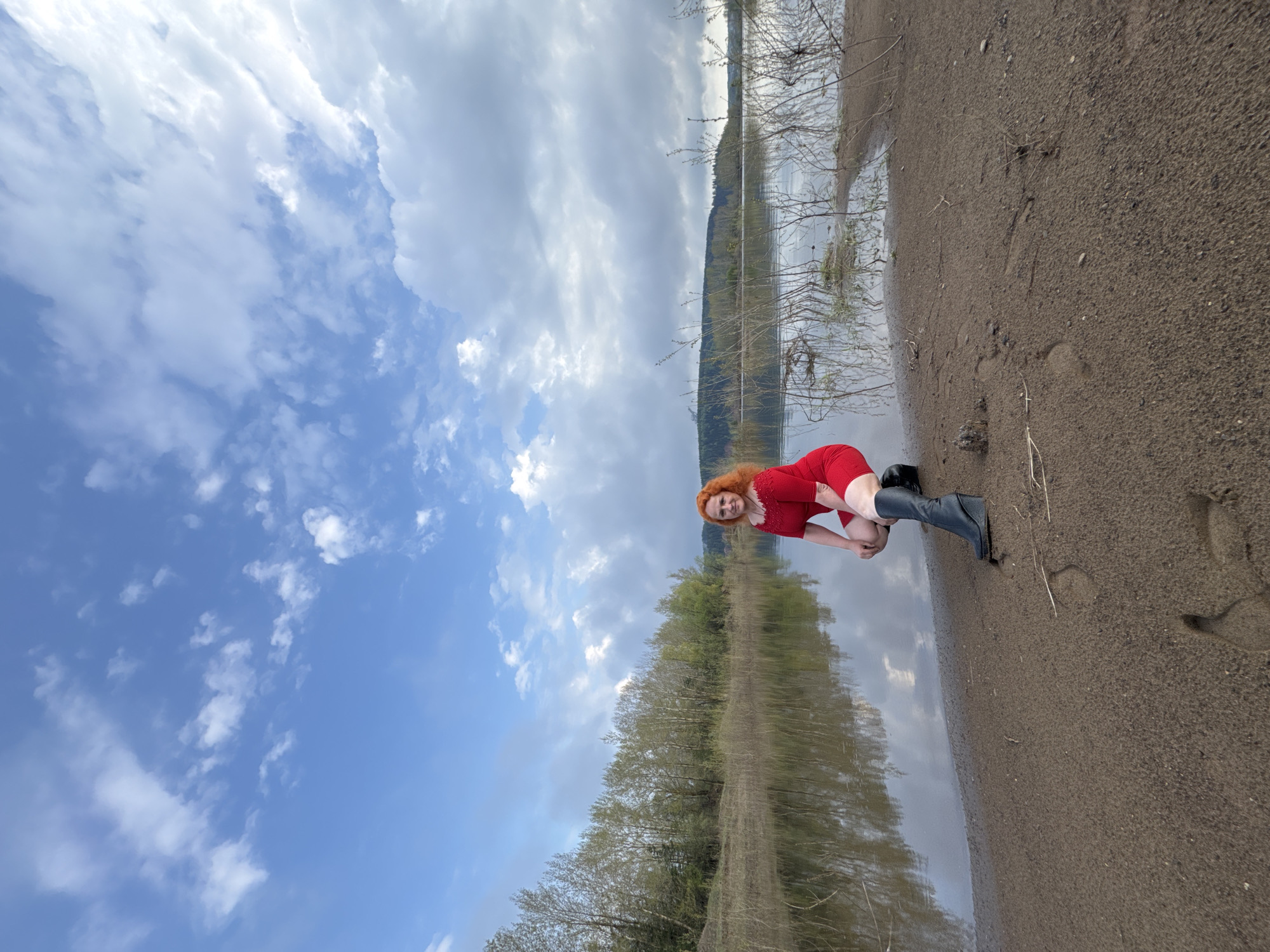 A woman with red hair wearing a red dress and black boots is kneeling on a sandy beach next to a body of water. The background shows trees reflected in the water and a cloudy sky. The location is unspecified, and there is no mention of Noviy, Russia in the provided information.