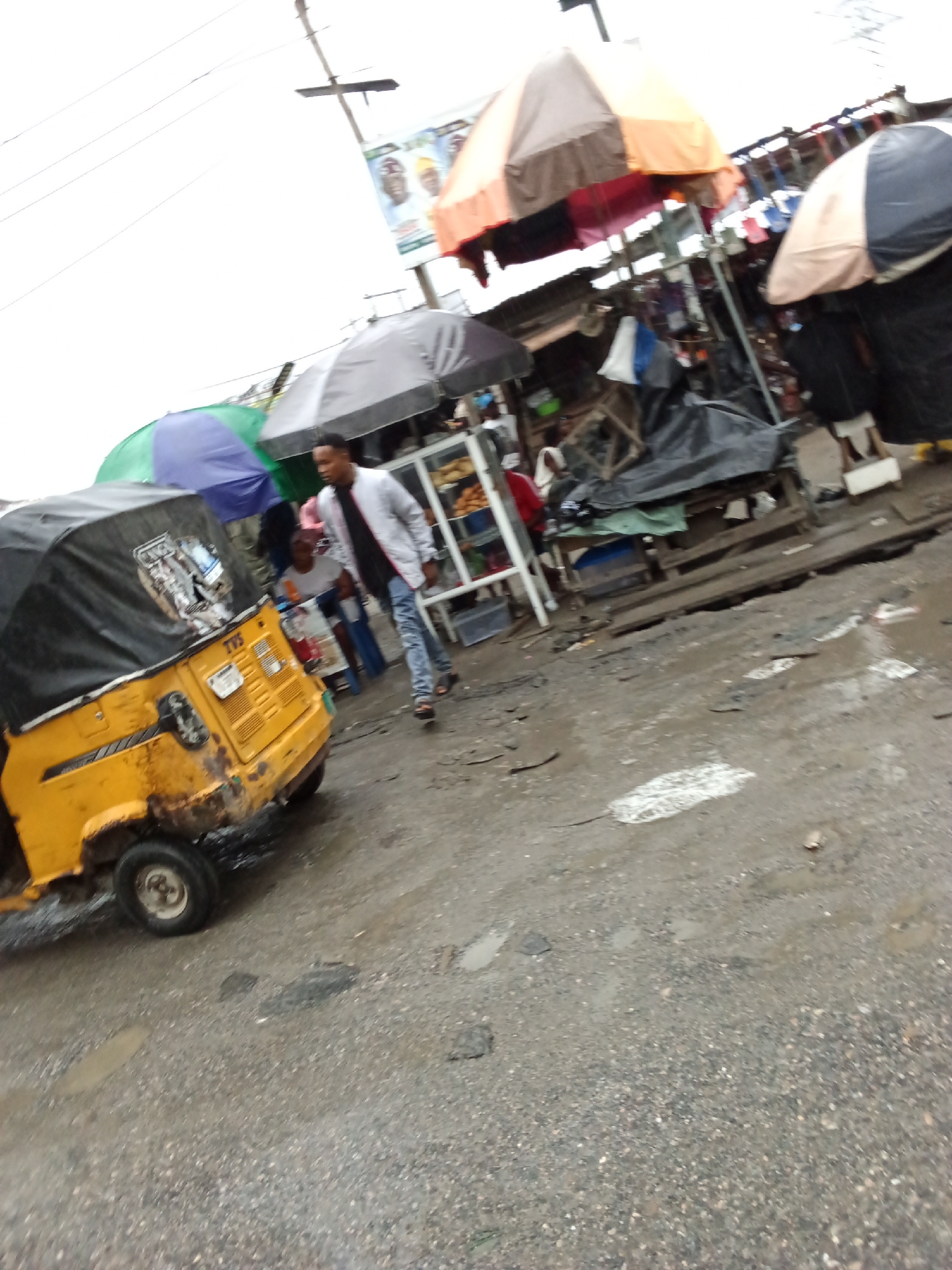 In an outdoor market setting in an unknown city, a yellow auto-rickshaw is parked on a wet, pebbled street. The rickshaw is covered with a black tarp. A young man in a light-colored jacket and blue jeans is walking away from the rickshaw. Behind him, there are market stalls with colorful umbrellas, and various items are displayed. In the background, banners with images of men are visible. The sky is overcast.