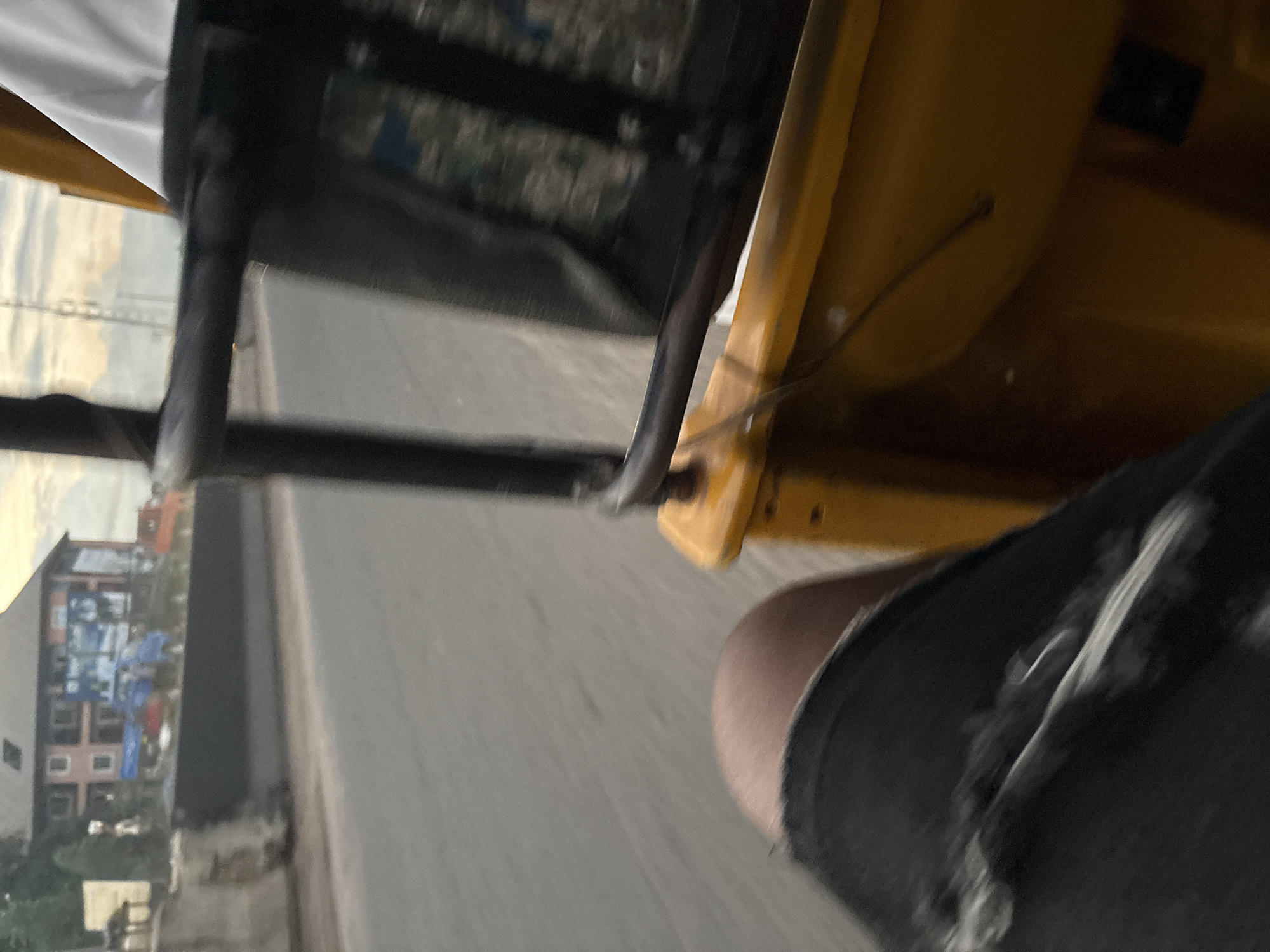 This is a first-person perspective shot from inside a yellow auto-rickshaw in Onitsha, Nigeria, likely on a street with moderate traffic. The foreground is dominated by the right leg of the passenger, wearing distressed denim shorts. To the right, a portion of the auto-rickshaw's yellow interior and a black metal bar are visible. The middle ground shows a grey road with tire marks and a concrete barrier. Beyond the barrier, there's a blurry view of a multi-story building with a red facade and blue banners on its side, possibly indicating advertisements. Several vehicles, including a blue truck and an orange truck, are partially visible in the background. The sky is overcast with hints of sunlight breaking through. The overall impression is of a typical urban commute scene in Onitsha.