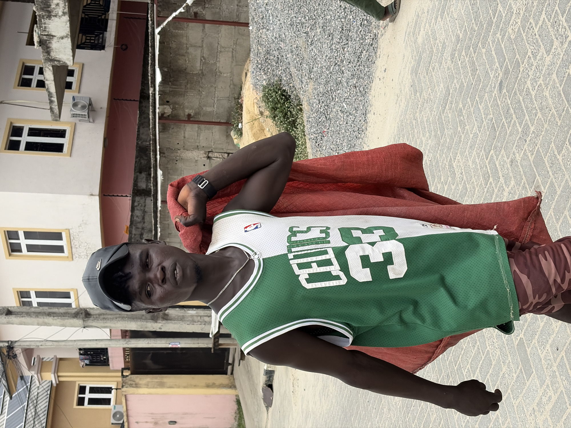 A man stands outdoors in Igboefon, Nigeria, wearing a black Nike baseball cap, a green and white Boston Celtics #33 basketball jersey, and maroon camouflage shorts. He has a red sack slung over his left shoulder, and a black watch is visible on his left wrist. He has a silver chain around his neck. The background includes a building with white framed windows, an air conditioning unit mounted on the wall, a gravel pile, and a patterned concrete walkway.