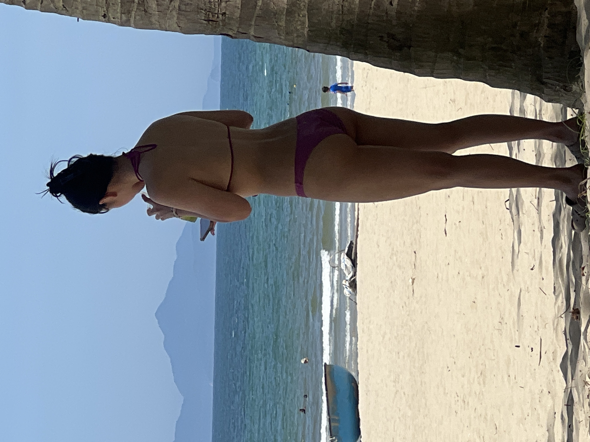 This image depicts a beach scene on a clear, sunny day. In the foreground, a woman in a purple bikini stands facing away from the camera, holding a drink in her right hand and a phone in her left. A palm tree trunk is to her right. The beach is sandy, with a few pieces of debris. In the midground, the ocean is visible with small waves. There is a blue boat on the left and a piece of white material on the sand to the right of the boat. Several people are in the water, and a young child in a blue swimsuit stands near the water's edge. In the background, mountains can be seen in the distance under a clear blue sky. This is likely a scene from one of Đà Nẵng's popular beaches, such as My Khe Beach or Non Nuoc Beach, known for their sandy shores, clear waters, and backdrop of mountains.