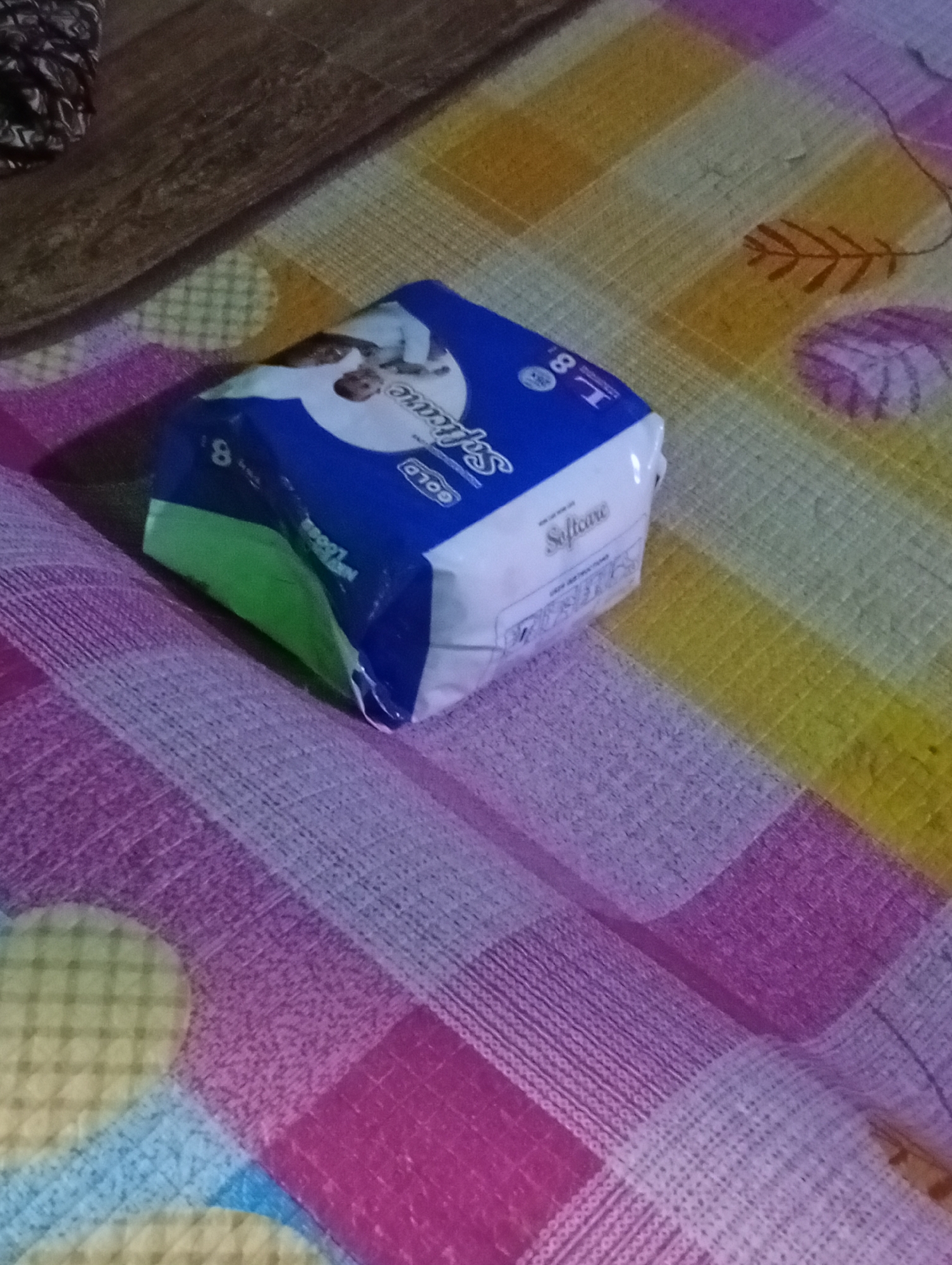 A package of baby diapers is lying on a patterned surface. The package is blue and white and has text and images on it. The surface appears to be a bedspread or blanket, with a repeating pattern of squares and lines in various colors, including pink, purple, yellow, and green. The lighting in the image is dim, suggesting it was taken indoors in low light conditions. The overall scene is unremarkable and does not provide any specific information about the city of Jalingo, Nigeria.