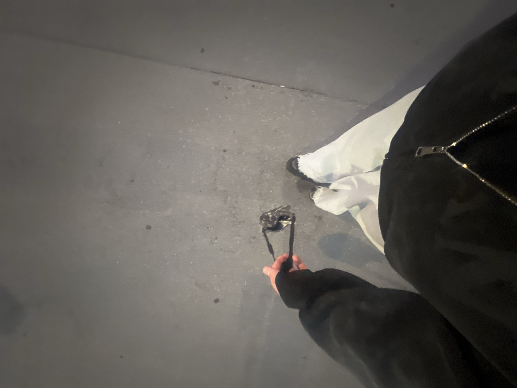 The scene is located in Saint Petersburg, Russia. The image is captured from a first-person, downward-looking perspective. A person's left arm and hand are visible in the left foreground, extended forward and holding a pair of folded dark-framed sunglasses with dark lenses. The person is wearing a black long-sleeved top or jacket, featuring a prominent silver zipper on the lower front. White trousers or pants are visible on the lower body, along with a black shoe on the left foot. The ground surface is a dark grey, textured material, consistent with asphalt or concrete, displaying various small specks and minor irregularities. A faint linear division or seam is present in the pavement extending diagonally across the lower right portion of the image. The environment is dimly lit.