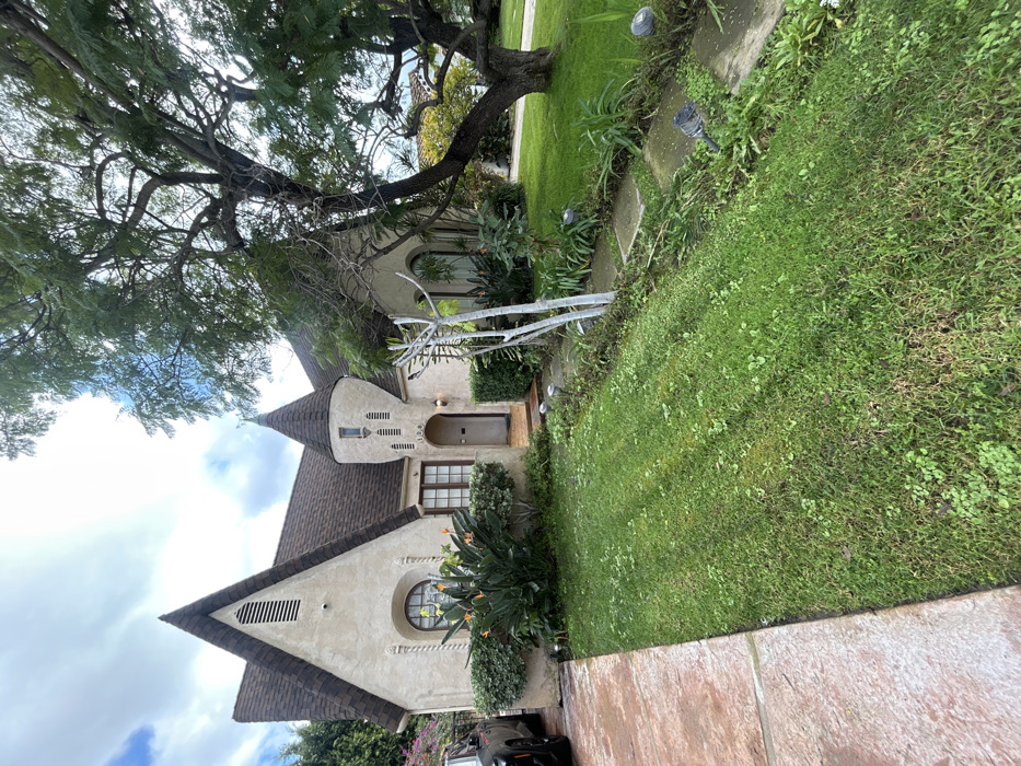 A fairytale-style house with a conical tower sits beneath a large tree. A dark roof, beige walls, and arched windows lend character. Lush greenery surrounds the house, with flowers blooming near the base. A green lawn stretches beside the house. Paving tiles are in the foreground. The sky is partly cloudy.