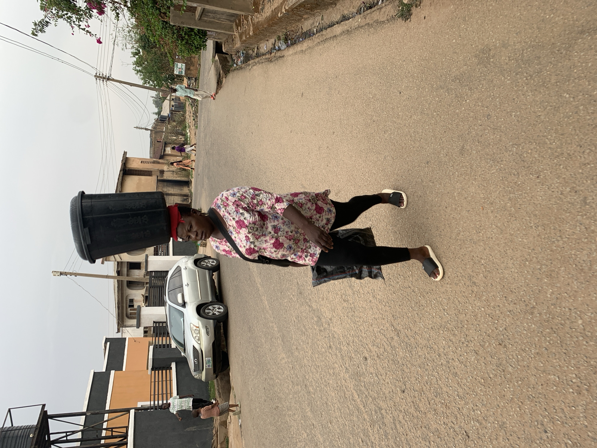 A woman balances a large, black plastic bucket on her head as she walks along a paved road in a residential area of Jos, Nigeria. She wears a floral-patterned blouse, black leggings, and black and white sandals. She carries a bag across her body. Behind her, a silver Lexus SUV is parked beside a modern building with a dark gray and tan facade. A black water tank stands on the street corner. People can be seen walking in the background. Utility poles and wires run along the street, and foliage is visible. The sky is overcast.