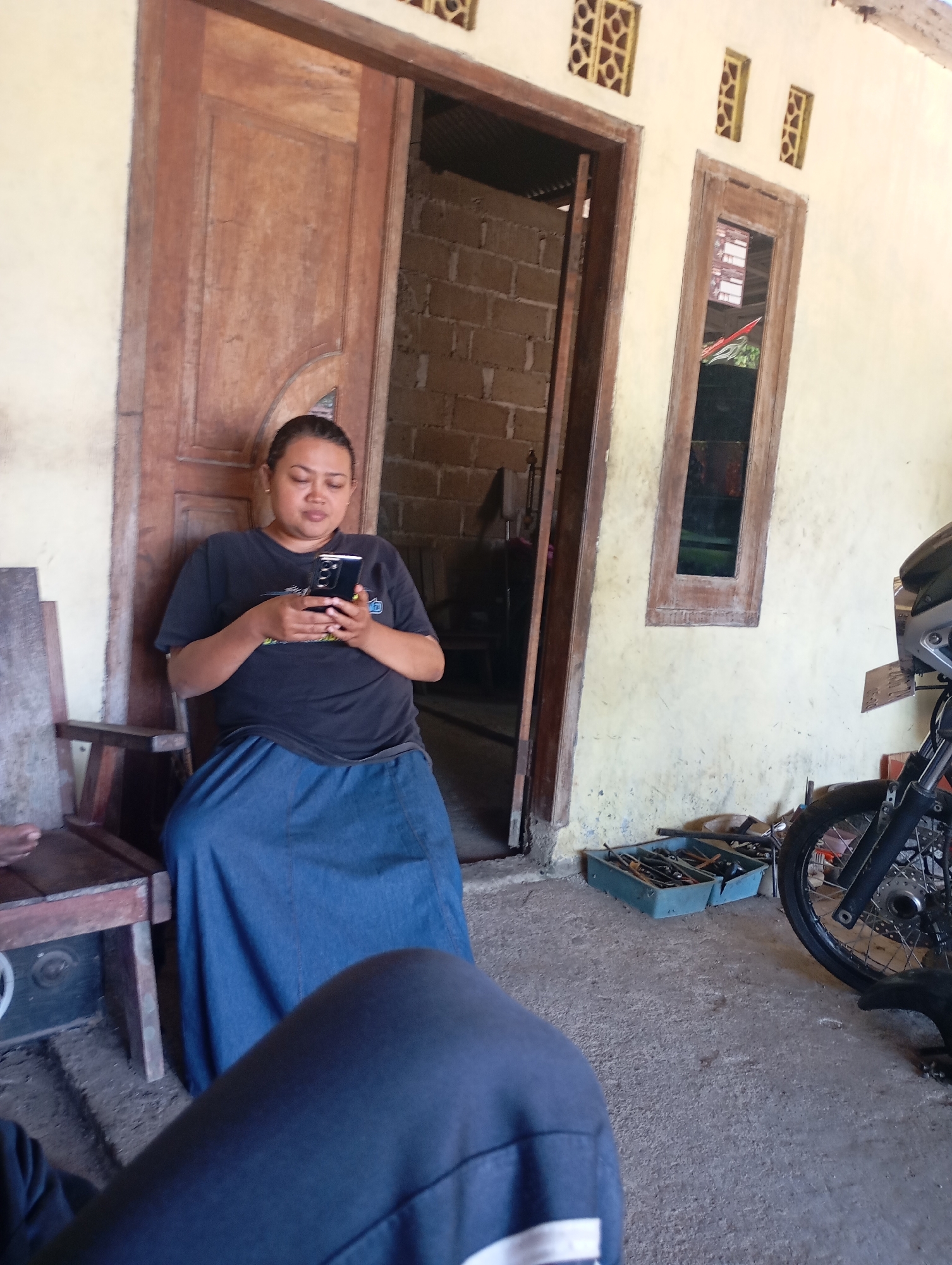 A woman is sitting on a wooden chair in front of a wooden door and a yellow wall, holding a smartphone and looking at it. She is wearing a dark t-shirt and a long blue skirt. Beside her is a wooden frame with a black seat. To her right, there is a window with a wooden frame and a motorbike wheel. On the ground, there is a blue toolbox with tools. The scene appears to be outdoors, possibly in a residential area in Ketepung, Indonesia, given the architecture and the presence of a motorbike. The ground is concrete and dirt.