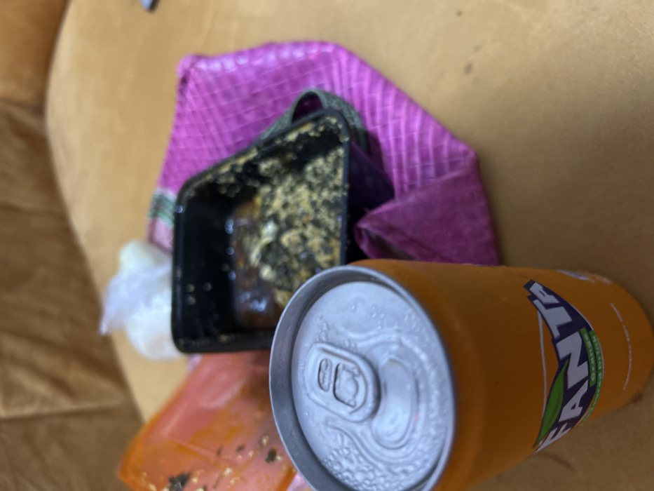 The image displays a close-up, elevated view of food and beverage items situated on a light brown, possibly wooden or upholstered, surface in Ilishan, Nigeria. In the immediate foreground, an upright, orange Fanta brand aluminum can is visible, covered in condensation. Its top, including the unopened pull-tab, is clearly detailed. Behind the Fanta can, towards the right, a black rectangular plastic food container holds partially consumed food consisting of a dark, viscous sauce or stew with lighter, granular solid components mixed within. This container rests upon a fuchsia-pink woven mat with a distinct striped pattern. To the left of the black container, an orange or reddish plastic item, possibly another container or bag, is present. Further back and slightly left of the black container, a small, clear plastic bag contains a white, amorphous substance. The background is out of focus. No individuals are depicted in the scene.