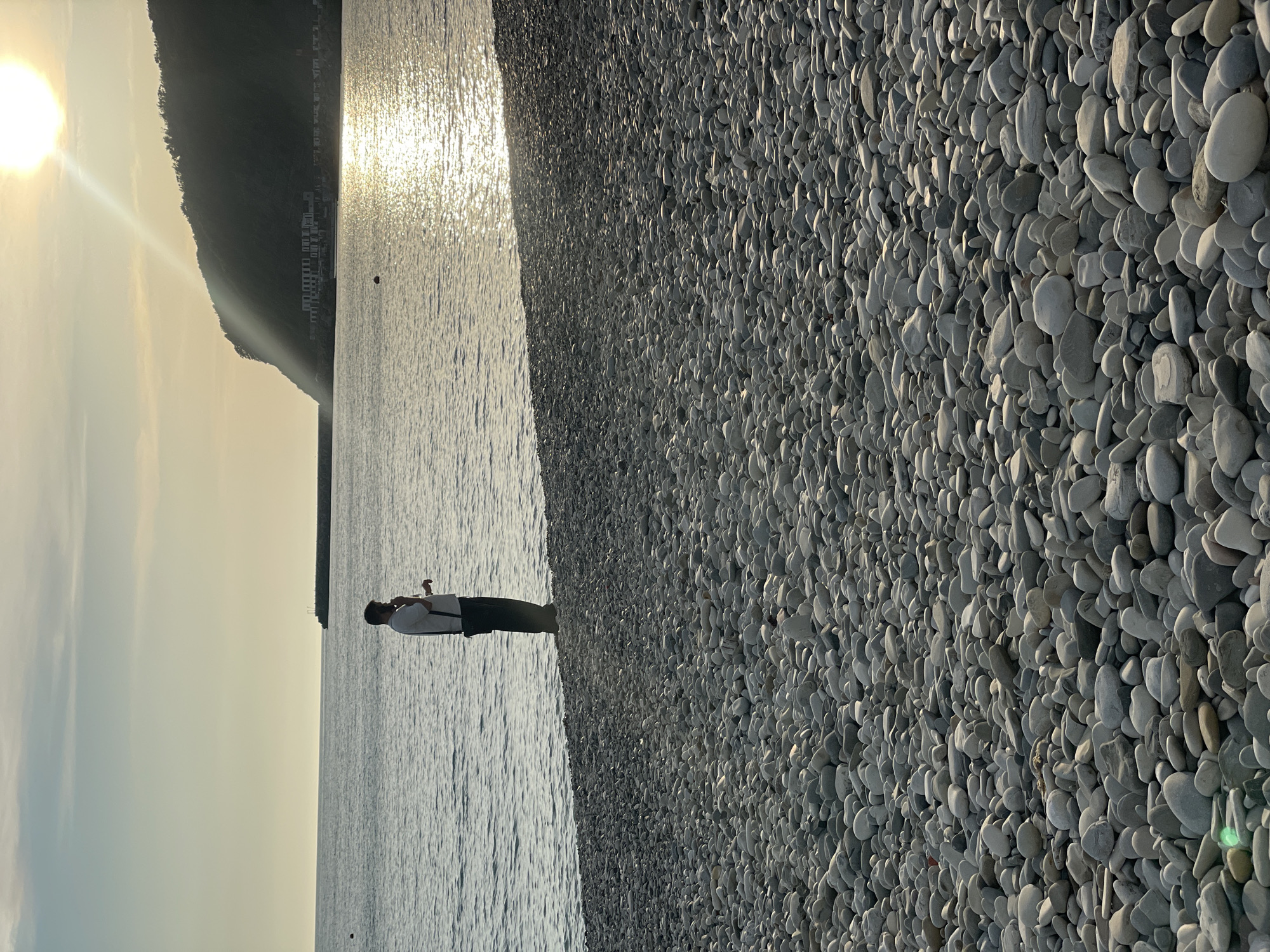 The photograph depicts a person standing in the water on a pebble beach in Olginka, Russia. The sun is shining brightly in the sky, reflecting on the water's surface. A large mountain is in the background. The beach is covered in smooth, gray pebbles. The person in the water appears to be holding something and looking at it.