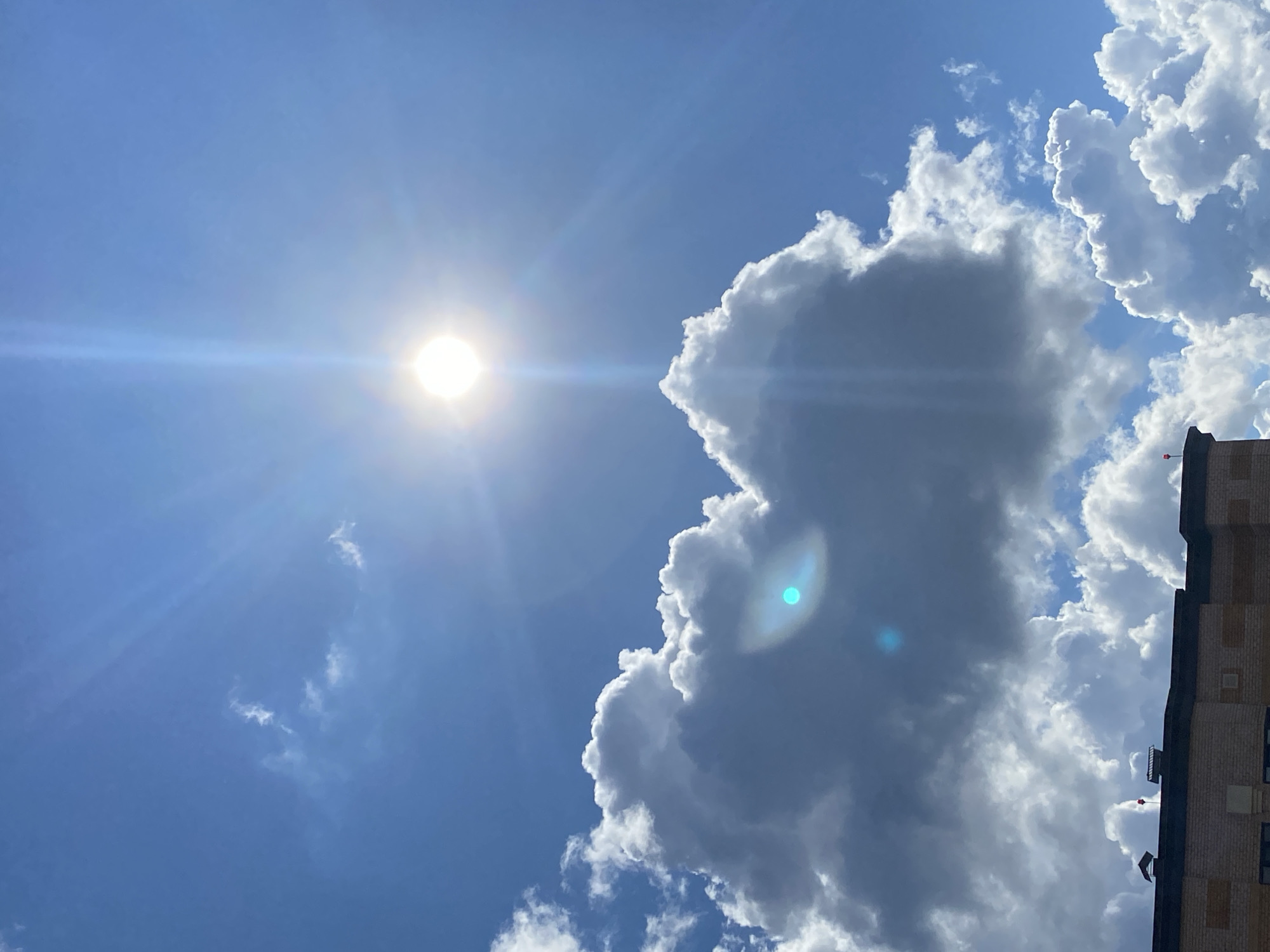 A bright, radiant sun shines directly overhead, its light casting strong rays downwards. Below, a building with a brick facade and dark roofing is visible. Fluffy, white cumulus clouds dominate the sky, with some areas appearing darker and more dense. A distinct lens flare is visible on one of the larger clouds, appearing as a bright blue-green halo. The overall scene suggests a clear, sunny day.