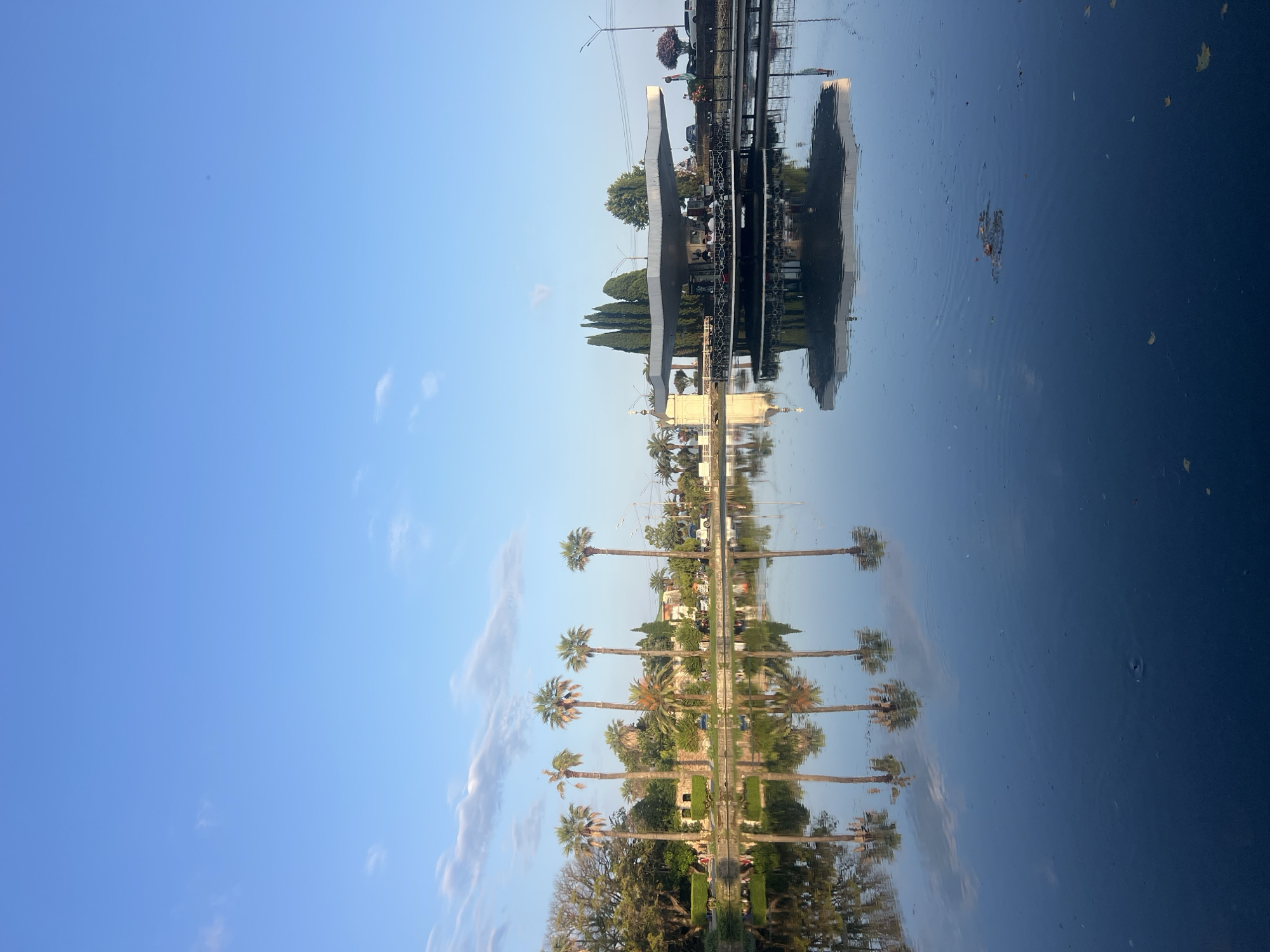 The image captures a serene waterscape with reflections of palm trees and a unique architectural structure in the city of New Athos, Abkhazia. A calm, dark body of water dominates the foreground, mirroring the clear blue sky and wispy clouds above. The reflections are sharp, showing several tall palm trees lining the shore on the left, their fronds creating intricate patterns. Further to the right, a distinctive modern structure with a wide, flat, angular roof stands over the water, supported by pillars. This structure appears to be a cafe or restaurant, with small figures visible sitting at tables on a lower deck. The architectural style is modern and somewhat geometric. Behind the palm trees, there are buildings and more vegetation, including cypress trees near the modern structure. The overall atmosphere is tranquil and picturesque, suggesting a pleasant day in a coastal or lakeside resort town.