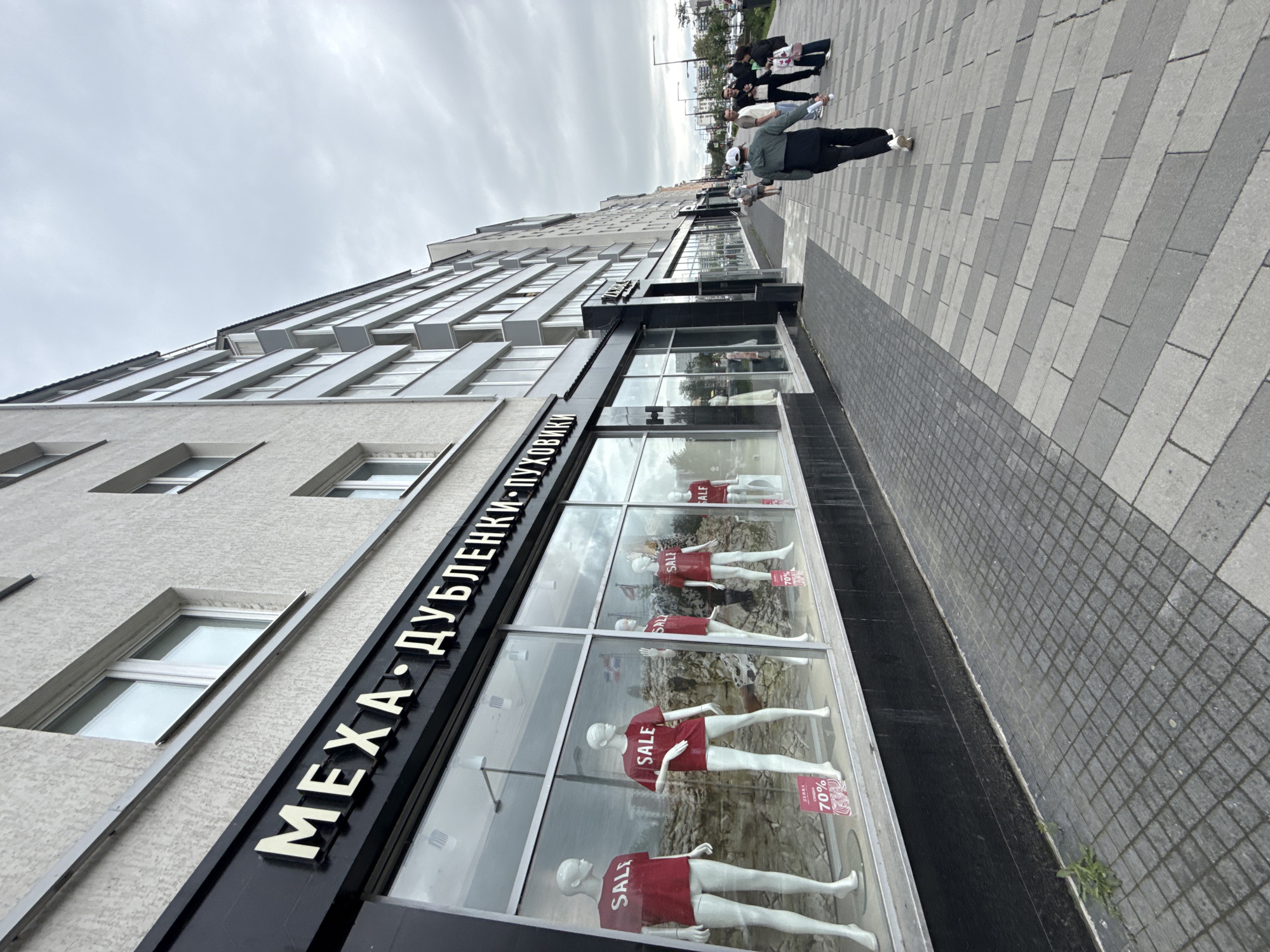 The image shows a street view of a commercial building with a large storefront. The storefront has mannequins displayed inside, wearing red shirts with "SALE" written on them. Some of the signs also indicate "70%". The building has multiple windows and a sign in Cyrillic script that appears to read "МЕХА • ДУБЛЕНКИ-ПУХОВИКИ" which translates to "FURS • SHEEPSKINS-DOWN JACKETS".
Several people are walking on the sidewalk in front of the store. The sky is overcast, suggesting it is daytime and the weather is cloudy. The sidewalk is paved with grey and lighter grey rectangular tiles laid in a diagonal pattern. The building itself has a modern architectural style with angled window frames. The overall scene depicts a commercial street in what appears to be an urban environment, possibly in Perm, Russia, given the Cyrillic text. The activity is simply people walking by and shopping. There are no strong emotional cues visible.