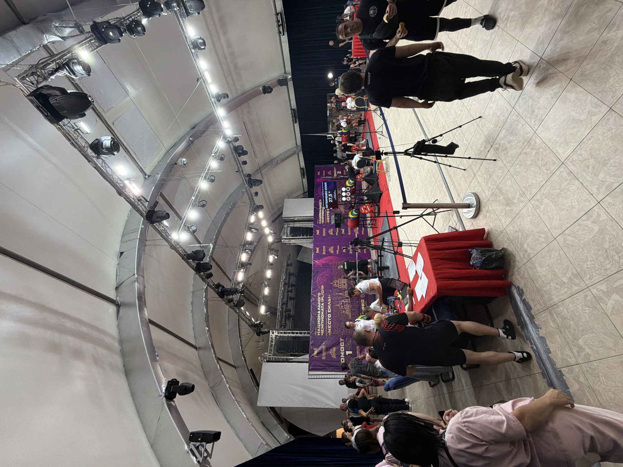 The image captures an indoor sporting event, likely a powerlifting competition, taking place within a large, tent-like structure. In the background, a stage is set up with a large purple banner displaying Cyrillic text, including "НАЦИОНАЛЬНОГО ЧЕМПИОНАТА IPL/СПР" (National Championship IPL/SPR) and "МЕСТО СИЛЫ" (Place of Strength). The stage is illuminated by numerous spotlights.
In the foreground and midground, a crowd of people are gathered. Some are spectators, while others appear to be athletes or event staff. A red-draped table is set up with papers and a laptop, suggesting an information or registration desk. Several tripods with cameras are positioned around the area, indicating filming or photography of the event. The floor is tiled, and a blue rope barrier separates different sections of the venue.
In the mid-left of the image, weightlifting equipment, including colored weight plates, is visible on a red carpeted platform. People are actively engaged in the competition or in discussions related to it. The overall atmosphere suggests an organized sporting event. The event is located in Suzdal, Russia.