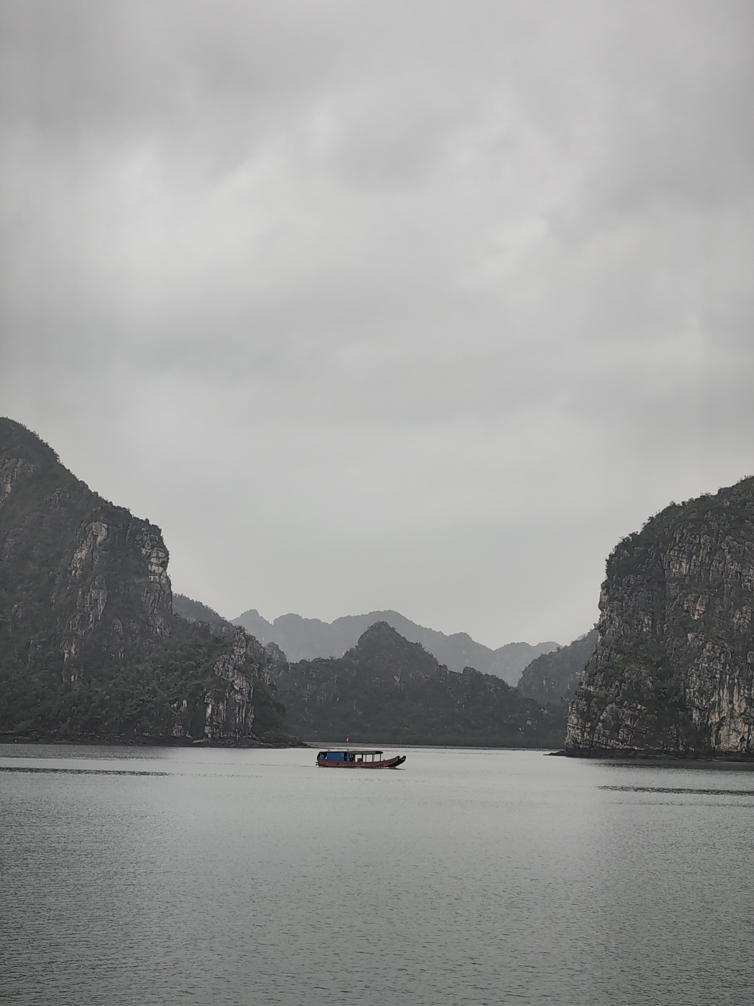 The image shows a boat on the water, surrounded by limestone cliffs under an overcast sky in Bai Chay Ward, Vietnam. The cliffs are covered in vegetation. The water appears calm and gray. The boat has a red hull and blue cabin.