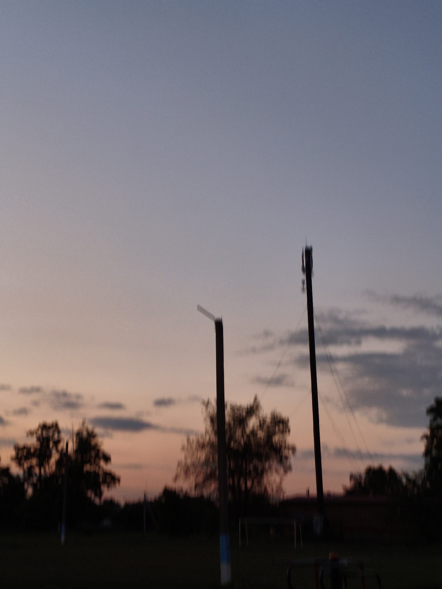 The image captures a dusk scene in Ustyvytsia, Ukraine. The sky is a gradient of pale blue at the top, transitioning to a soft orange and pink near the horizon, indicating the setting sun. Scattered clouds, dark grey and elongated, are visible against the lighter sky. In the foreground, silhouettes of trees with full foliage create a dark mass. Two tall, dark poles, one on the right with visible equipment near its top, stand prominently. There is also a shorter, dark pole in the center of the frame, partially obscured by a thin, lighter pole with a blue stripe at its base, and another similar pole to the left. In the distance, a dark, low building or structure is barely discernible behind the trees, with wires connecting to the poles. The ground is dark and appears to be a grassy area. The overall atmosphere is tranquil, characteristic of a rural evening.