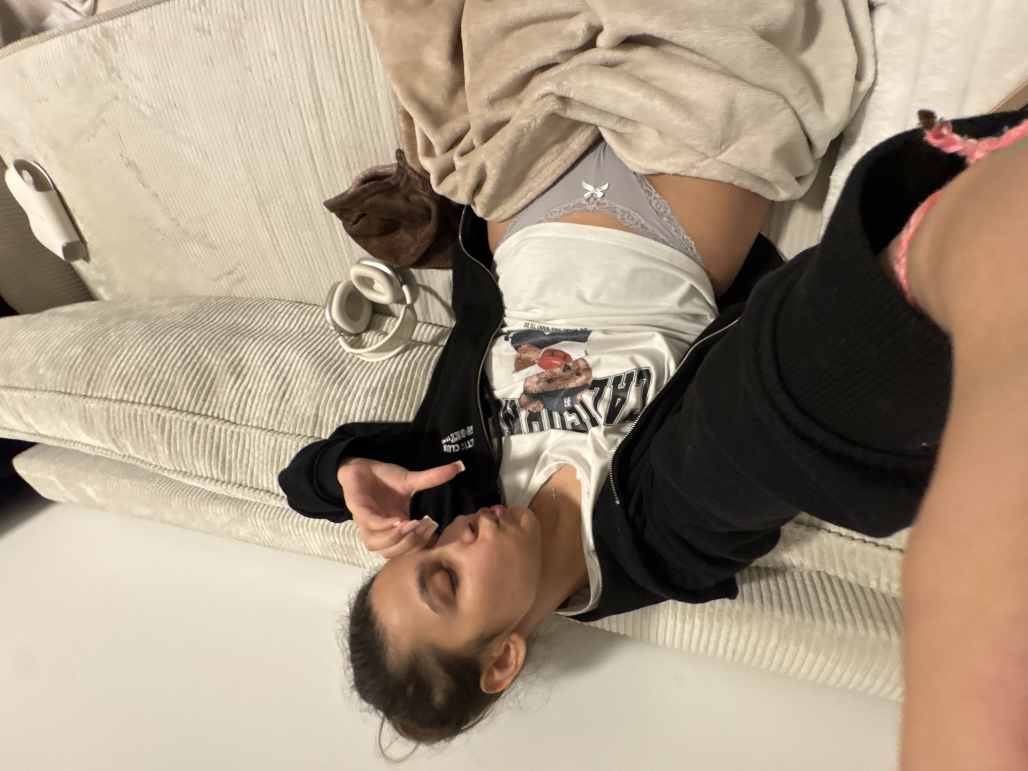 The image, taken in Mönchengladbach, Germany, features a person lying on a light-colored, corduroy-textured sofa. The person, likely female, has dark hair tied back and appears to be resting or sleeping, with closed eyes and one hand near her face. She is dressed in a white t-shirt, a black zipped hoodie, and light grey, lace-trimmed underwear adorned with a small white bow. A beige blanket is partially draped over her lower body.
On the white t-shirt, a graphic print depicts what appears to be a teddy bear or similar animal, with partially visible text including "DO WHAT YOU WANT TO DO" and "BEAR." A small, cross-shaped pendant is visible on her chest.
Next to the person on the sofa, a pair of white over-ear headphones and a small white object, possibly a remote or charging case, are placed. A brown fabric item is also seen nearby. The scene suggests an indoor, relaxed domestic setting, likely a living room. The photograph is oriented sideways, with the person's head positioned towards the lower left and her body extending diagonally upwards. The overall atmosphere conveys peace and comfort.