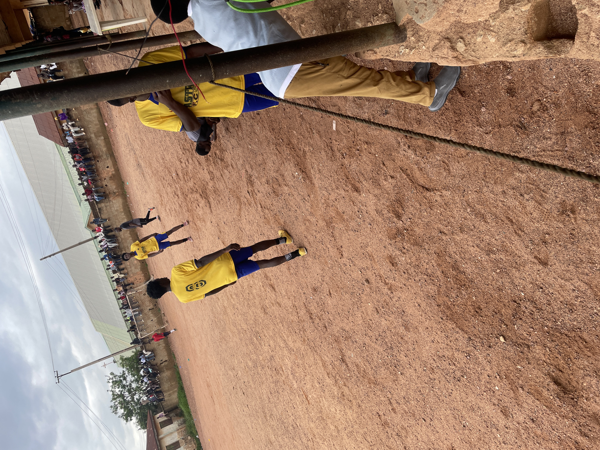 The image depicts an outdoor scene, likely a sports field, with a reddish-brown dirt surface. Several individuals are present, some of whom appear to be athletes in yellow shirts and blue shorts, and others spectators sitting on a raised wall in the background. One person in the foreground is wearing a light blue shirt and brown pants. A thick rope is visible running diagonally across the frame, secured to a rough, reddish-brown post on the right. The environment suggests a warm climate, with a cloudy sky overhead. The presence of a wall with people sitting on it and a goal net in the distance indicates a sporting event is either in progress or has recently concluded. The overall atmosphere is casual and appears to be a public gathering in an urban or semi-urban setting within Jos, Nigeria.