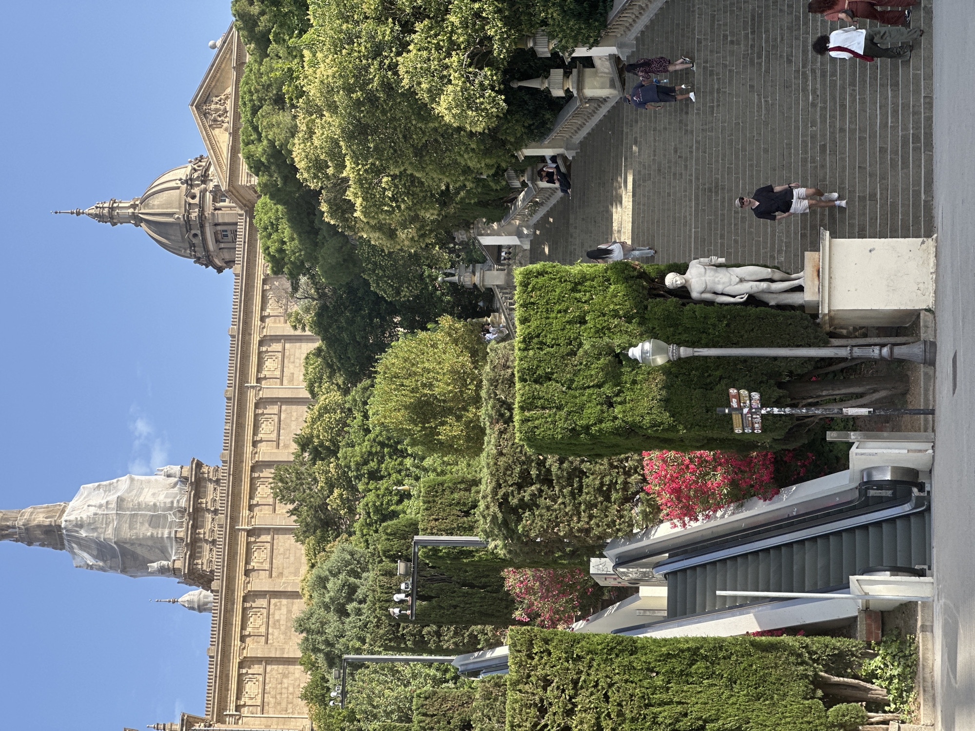 The image captures a section of the grand staircase leading up to the Palau Nacional on Montjuïc hill in Barcelona, Spain. The architectural style of the Palau Nacional, a large, imposing structure with a domed roof and classical detailing, is visible at the top of the scene. Lush greenery, including manicured hedges and mature trees, flanks the wide stone staircase and an adjacent escalator. Several people are ascending and descending the stairs, some walking individually, others in pairs or small groups. A statue of a male figure stands prominently near the base of the stairs. The sky is clear and blue, indicating a sunny day. The scene is characterized by its architectural grandeur, extensive landscaping, and pedestrian activity.