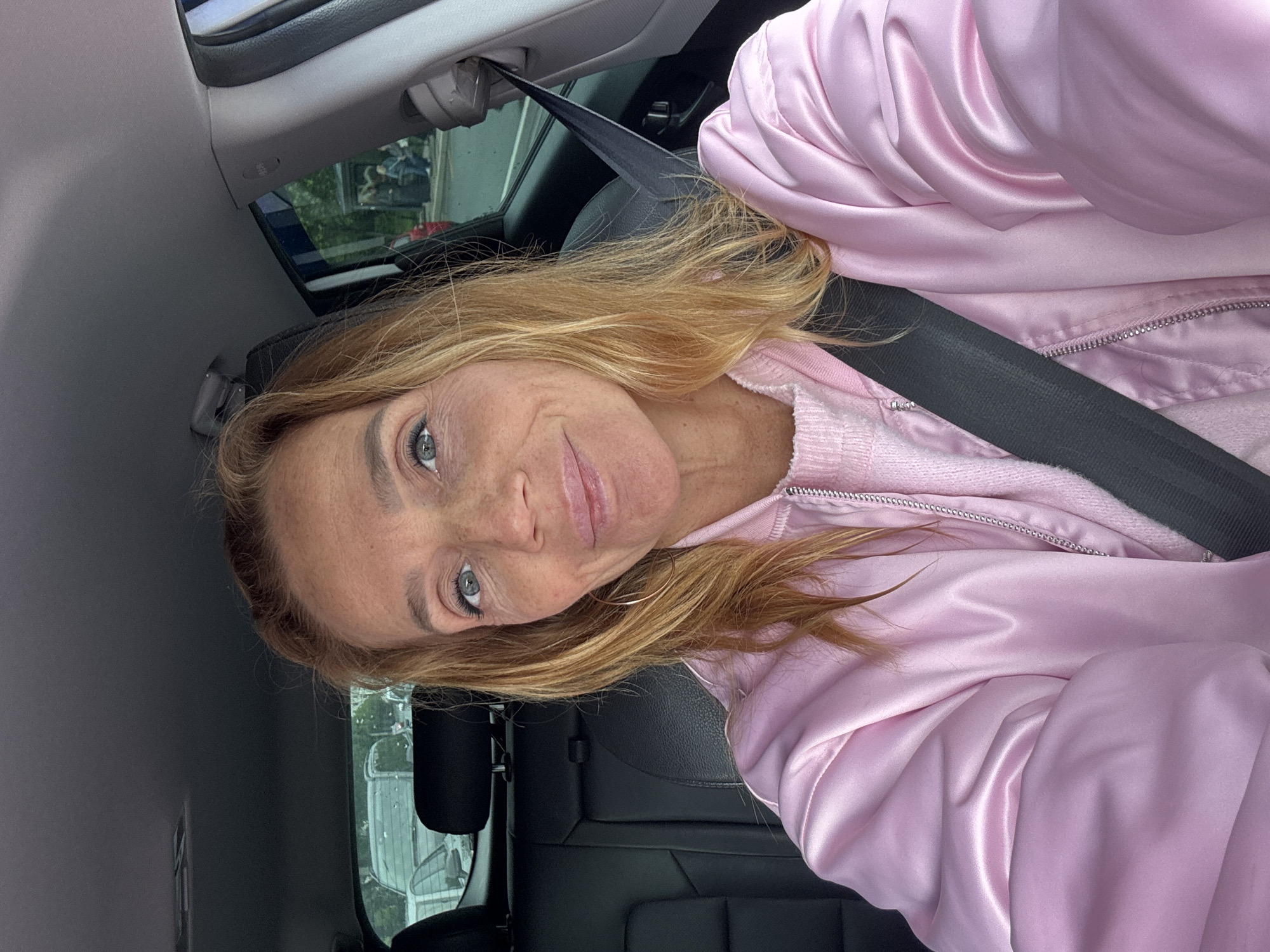 A person with long, wavy, blonde hair and gray eyes is seated in the driver's seat of a car, wearing a pink sweater and a shiny pink jacket. A black seatbelt is fastened across their chest. The interior of the car is visible, including the headrest, seat, and roof. Outside the car, a building and trees can be seen through the window, suggesting an urban environment. The location of the photo is unknown.